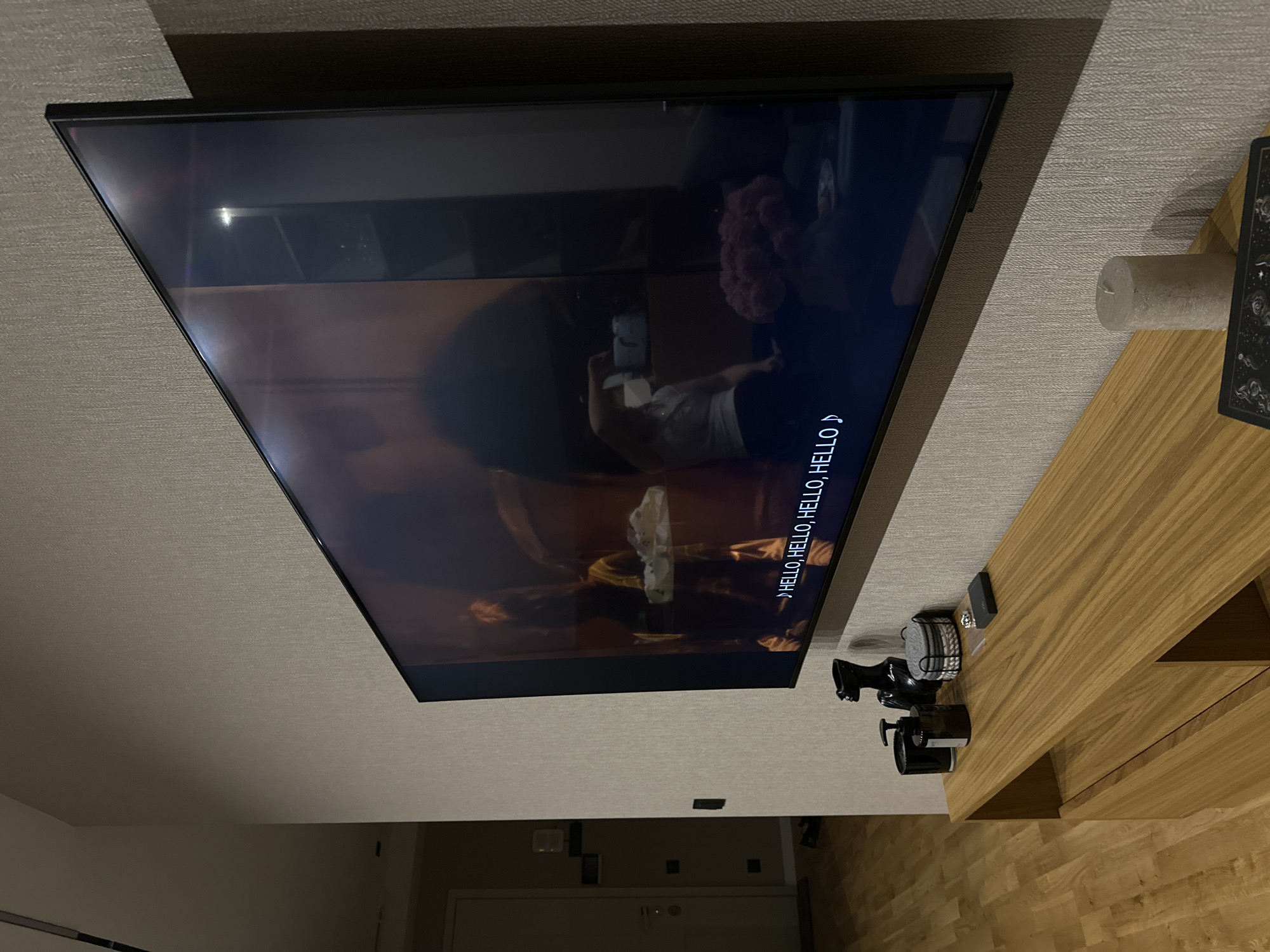 The image shows a television mounted on a wall. The television is displaying a scene with a person and the subtitles "HELLO, HELLO, HELLO, HELLO". A wooden table is to the right of the television. On the table are several items, including a candle, a deck of cards, and a set of black bottles and containers. The wall appears to be covered in a textured wallpaper. A door is visible in the background, suggesting the television is in a living room or similar space. The image doesn't provide enough information to pinpoint a specific location in Yerevan, Armenia.