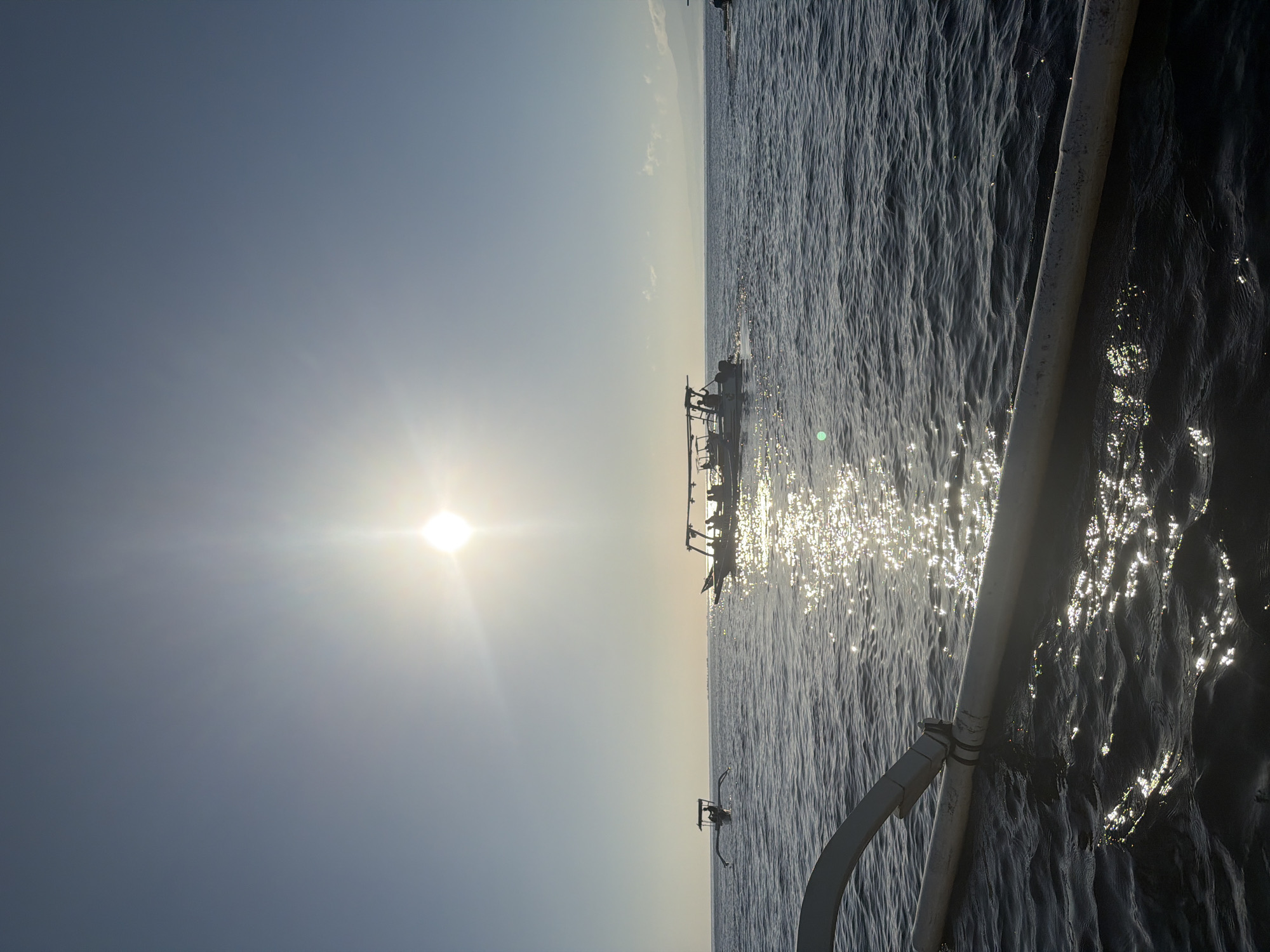 The image depicts a serene seascape with a bright sun low in the sky, casting a glittering reflection on the water. The viewpoint is from a boat, with the edge of its hull and outrigger visible in the foreground. In the middle ground, two traditional Balinese fishing boats, known as jukung, are present. The boat on the left is smaller and has a single outrigger, while the boat on the right is larger, with a canopy providing shade and several people visible on board. The boats are silhouetted against the bright sun. The background shows a hazy horizon with mountains faintly visible. The sea is calm, with gentle ripples. The overall impression is one of a tranquil morning or late afternoon at sea. The scene is likely captured in the waters off the coast of Seririt, Indonesia, a region known for its traditional fishing communities and serene coastal landscapes.