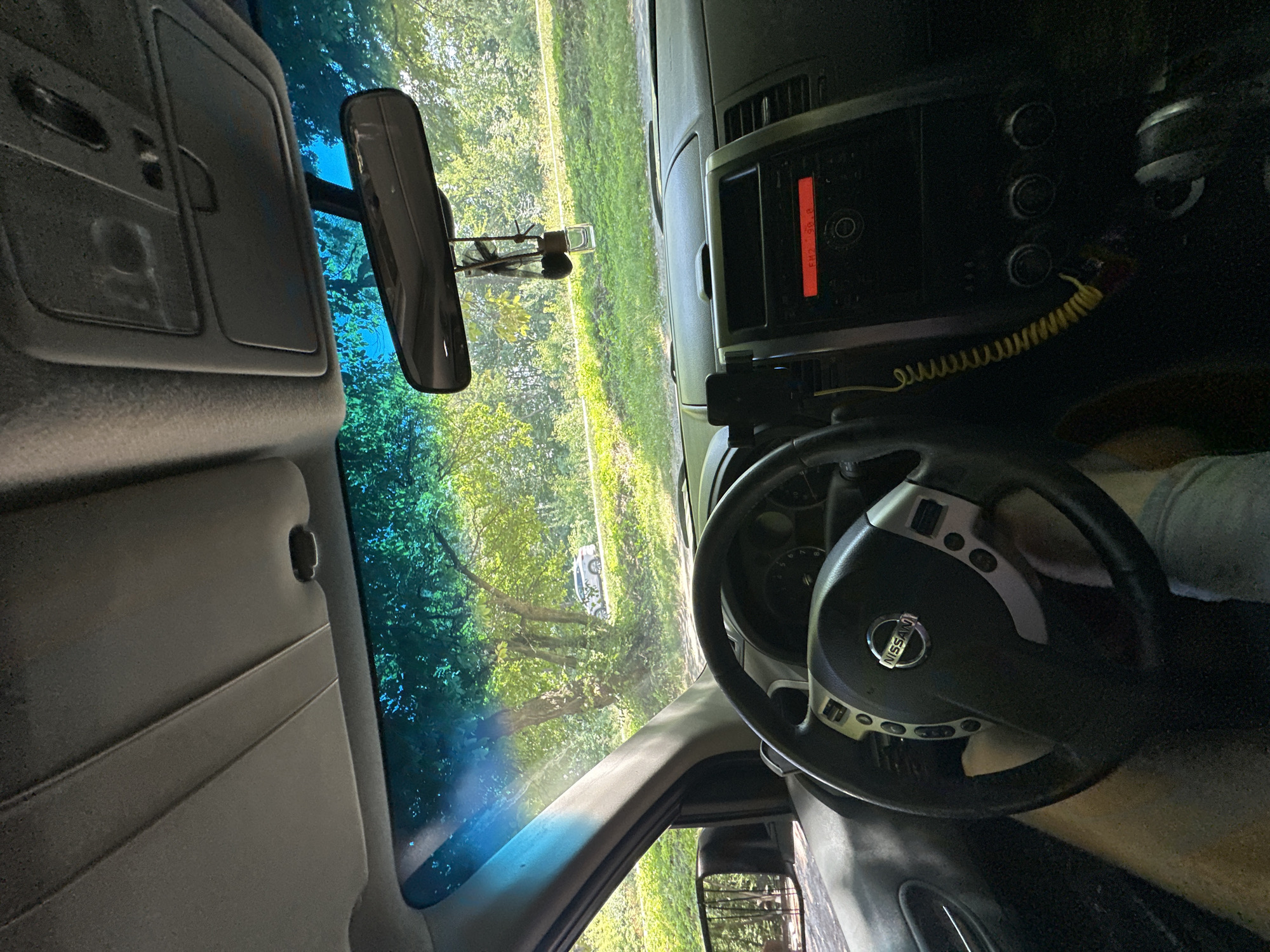 The image is a first-person perspective from inside a Nissan vehicle, parked on the side of a rural road. The interior of the car is visible, including the steering wheel with the Nissan logo, the dashboard with the radio displaying "FM2 99.8", a cell phone holder, and the gear shifter. A person's legs are visible in the driver's seat. Outside the windshield, a tree-lined road is visible, with a white SUV parked on the opposite side of the road. The scene appears to be in Shatava, Ukraine, based on the provided location context, suggesting a setting in a natural, possibly wooded, environment. The overall lighting suggests it is daytime.
