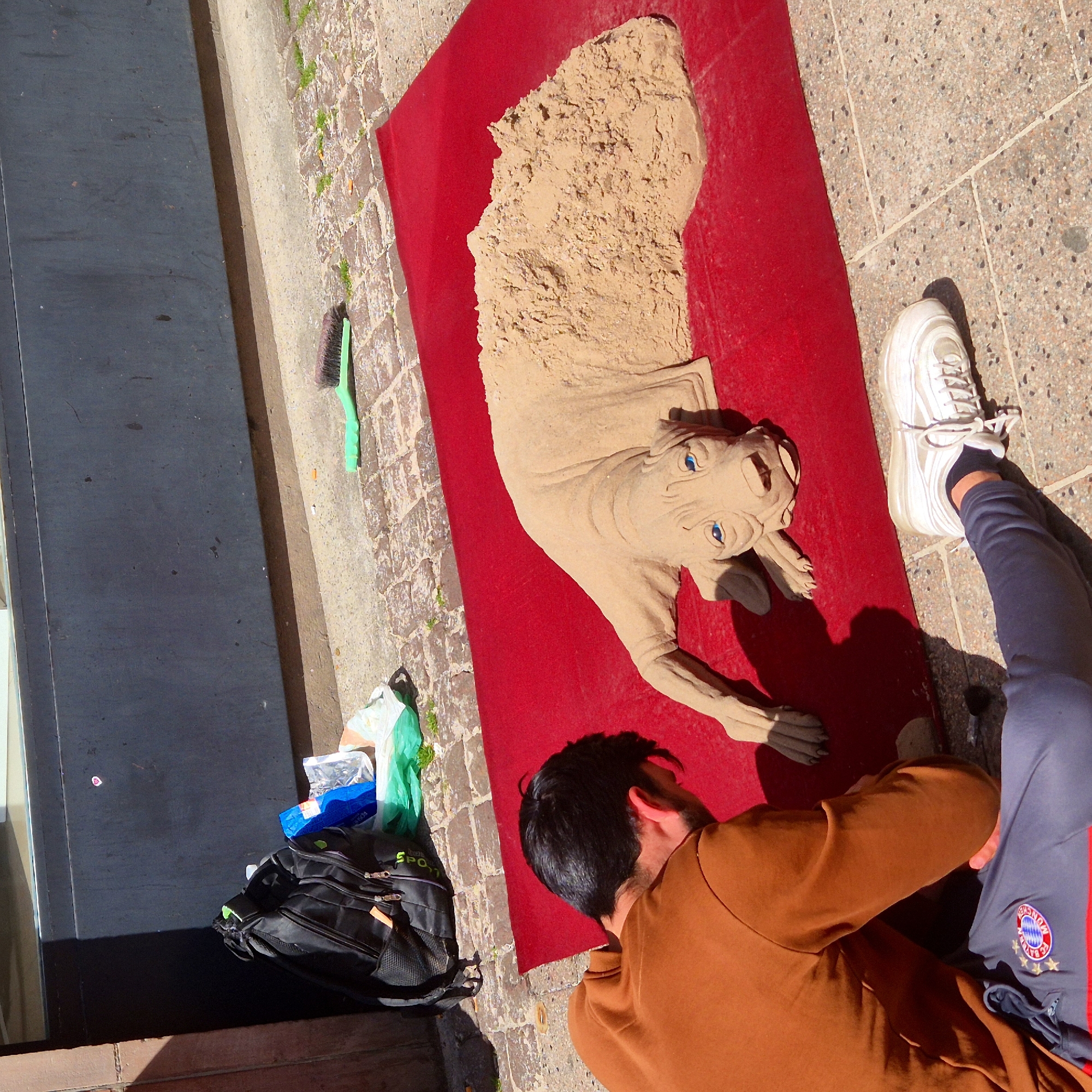 The image depicts a street scene in Strasbourg, France. A sand sculpture of E.T. rests on a red carpet laid out on the sidewalk. A man in a brown shirt and a person wearing grey athletic pants and white sneakers are nearby. To the left of the sculpture, a black backpack and a cleaning brush sit on the stone-paved sidewalk. The edge of a building is visible in the lower left. The sidewalk is paved with light grey cobblestones.