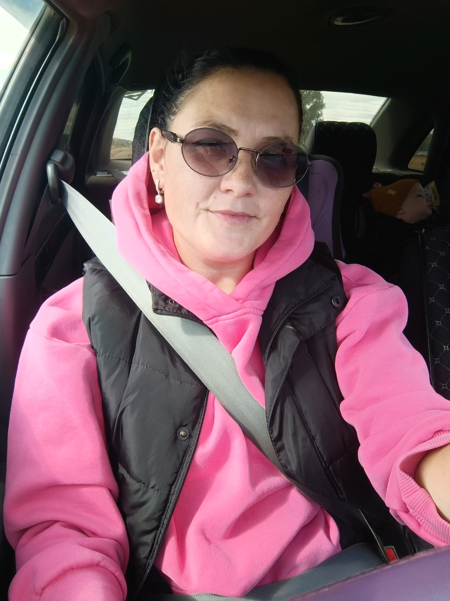 The image shows a woman with dark hair pulled back, wearing round sunglasses, a pink hooded sweatshirt, and a black puffer vest. A seatbelt is fastened across her chest. She is sitting in the driver's seat of a car, with the interior visible. A child's car seat is visible in the background, behind the woman. The environment outside the car appears to be a rural or undeveloped area, possibly with some trees. The lighting suggests it is daytime. The image does not provide any specific information about the location within the city of Chumna, Russia.