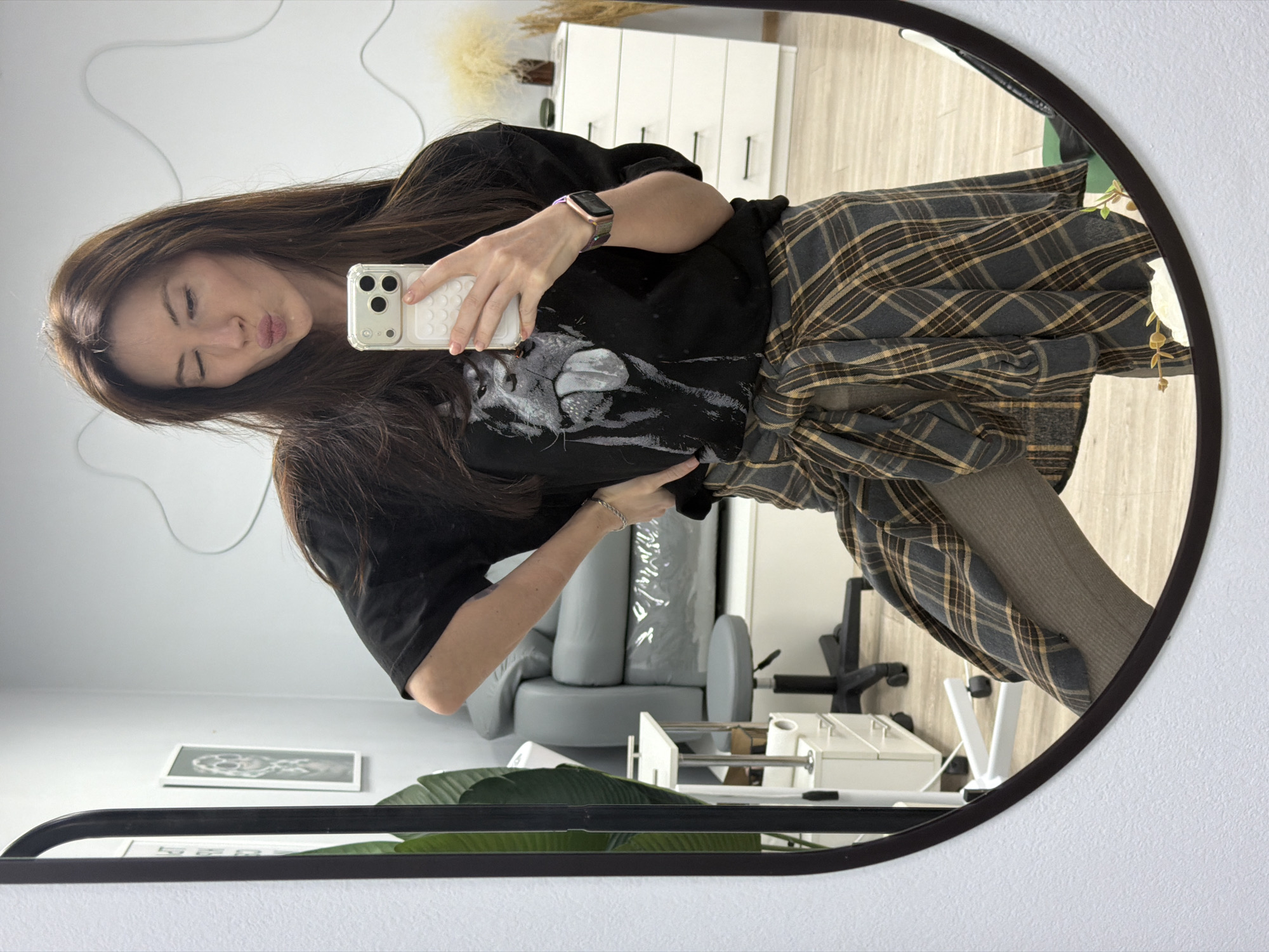 A woman is taking a selfie in a mirror, presumably in an indoor environment. She has long brown hair and is wearing a black t-shirt with a graphic print, and a plaid shirt tied around her waist. Her lips are puckered in a kissing expression. She is holding a white phone in her right hand to take the photo. The mirror is round with a dark frame and reflects a room with a white dresser, a gray couch, and a rolling chair. A black frame on the wall is visible in the bottom left corner of the mirror's reflection. There's a plant with large green leaves near the bottom of the image. The city and country of Perm, Russia, are not evident in this image.