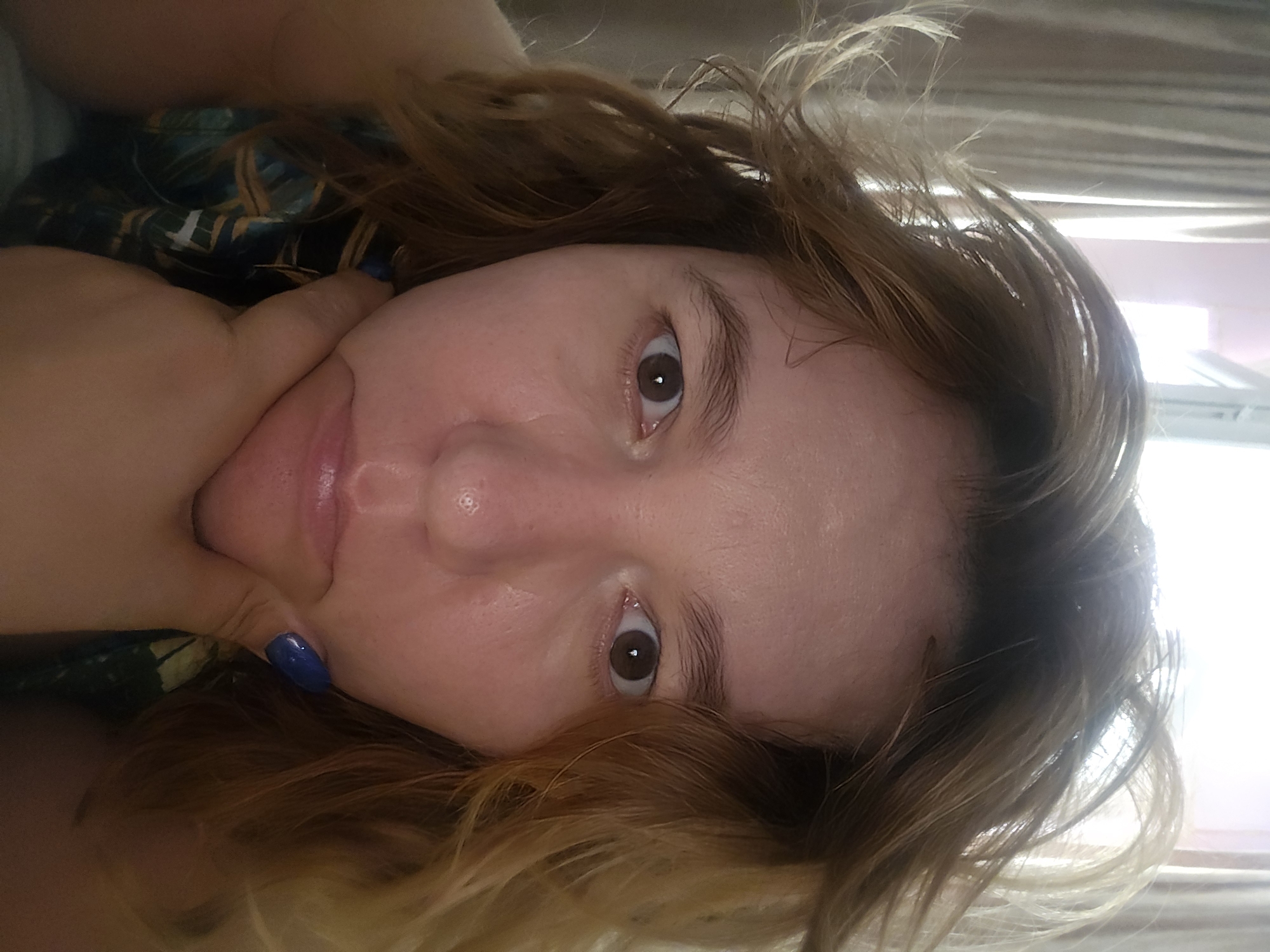A close-up, frontal, medium-shot photograph shows a woman in her late 20s to early 30s with light skin, light brown, tousled hair, and brown eyes. Her left hand is resting on her chin, and her right hand is not visible. She is wearing a dark top with a patterned design. The background is out of focus and shows a wall with curtains and a window. The lighting is bright and appears to be natural. The woman is looking directly at the camera with a neutral expression.