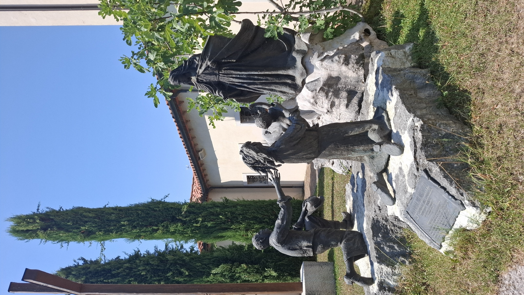 This image captures a bronze sculpture situated in a garden setting, with a building and cypress trees in the background under a clear blue sky. The sculpture depicts several figures. In the foreground, a kneeling male figure is extending his hand towards another kneeling figure. This second figure, also kneeling, appears to be a woman holding a child. Behind these figures, a larger, robed figure stands on a rock formation, with its arms extended. A wooden cross is visible on the left side of the image, partially obscured by a tree. A plaque is affixed to a rock in the foreground, providing inscription details. The surrounding environment includes green grass and a paved area. The sculpture is located in the city of Motta di Livenza, Italy, and specifically in front of the Church of San Giovanni Battista.