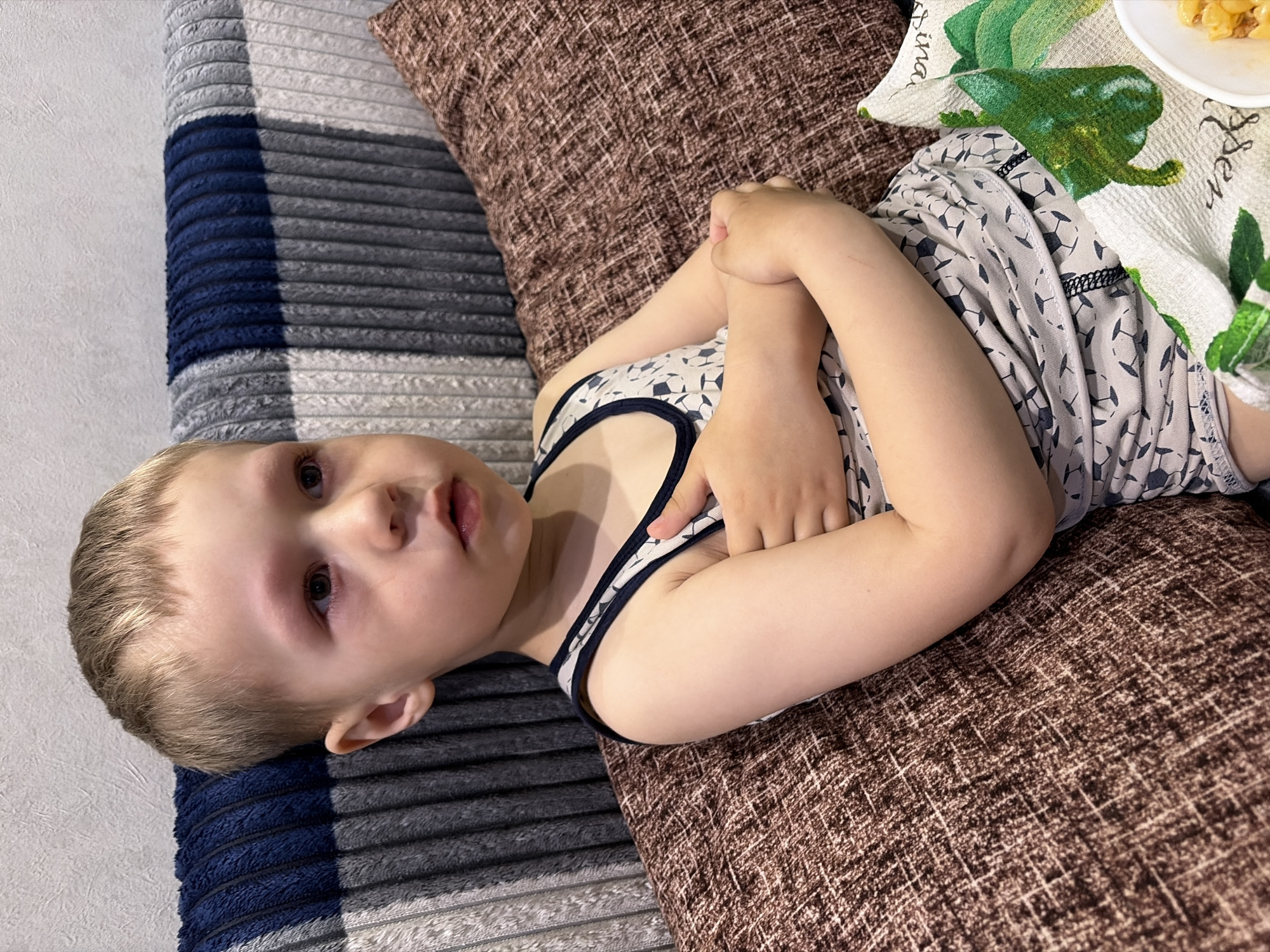A young child is lying on a textured brown pillow on what appears to be a striped bed or cushion. The child is wearing a patterned tank top. A patterned towel and a plate of food are partially visible in the top right corner of the image. The photo was taken indoors. There is no indication about the location.