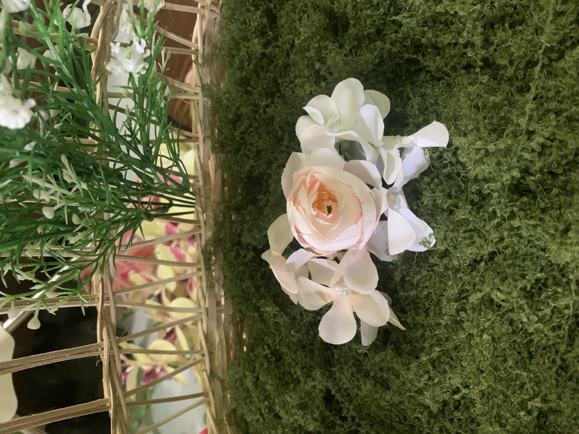 A close-up, overhead shot displays a delicate arrangement of artificial flowers placed on a bed of green moss. The flowers, predominantly white and a soft pink, are clustered together, with a central pink rose showing intricate petal detail. Behind the flowers, a woven basket structure made of light brown reeds is visible, with green artificial foliage emerging from it. The background is blurred, showing hints of more colorful floral arrangements. The scene appears to be an indoor decorative setting, possibly for an event or a craft project. There is no indication of a specific location within Kuala Pembuang, Indonesia.