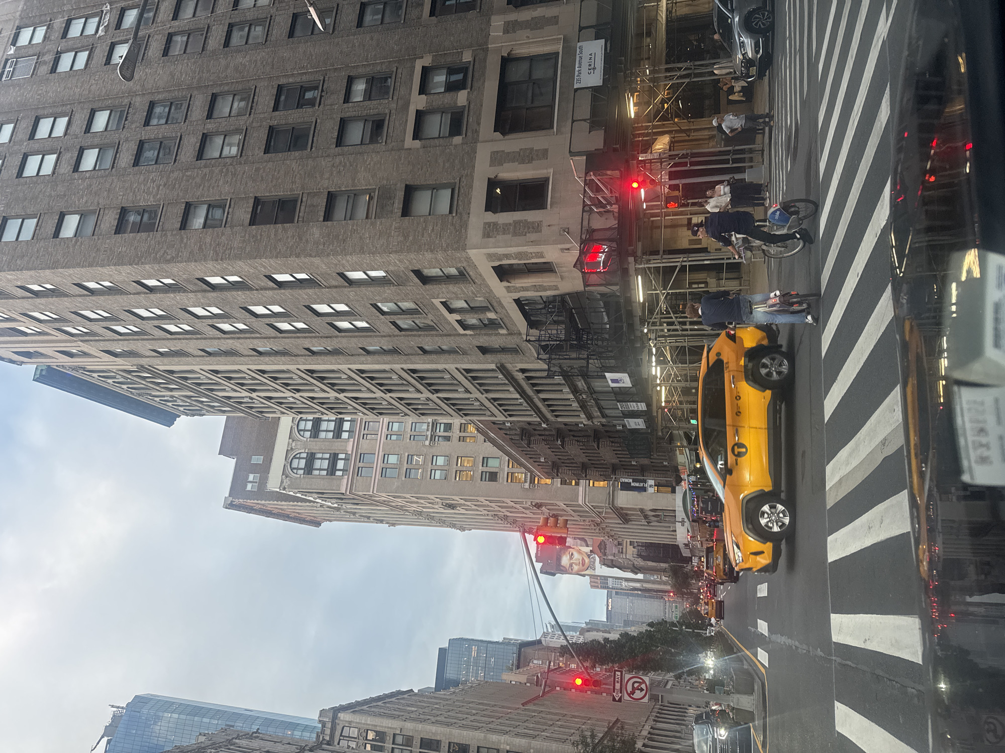The image captures a street scene in New York City, United States of America. A yellow taxi, a Toyota RAV4, is in motion, crossing a crosswalk. Two cyclists are also present, one on a pink bicycle and another on a blue Citi Bike. Pedestrians are visible on the sidewalks. The environment features tall buildings lining the street, a traffic light displaying red, and signage for "ONE WAY" and a no-U-turn symbol. The sky is overcast. The scene appears to be captured from inside a vehicle, looking forward through the windshield.