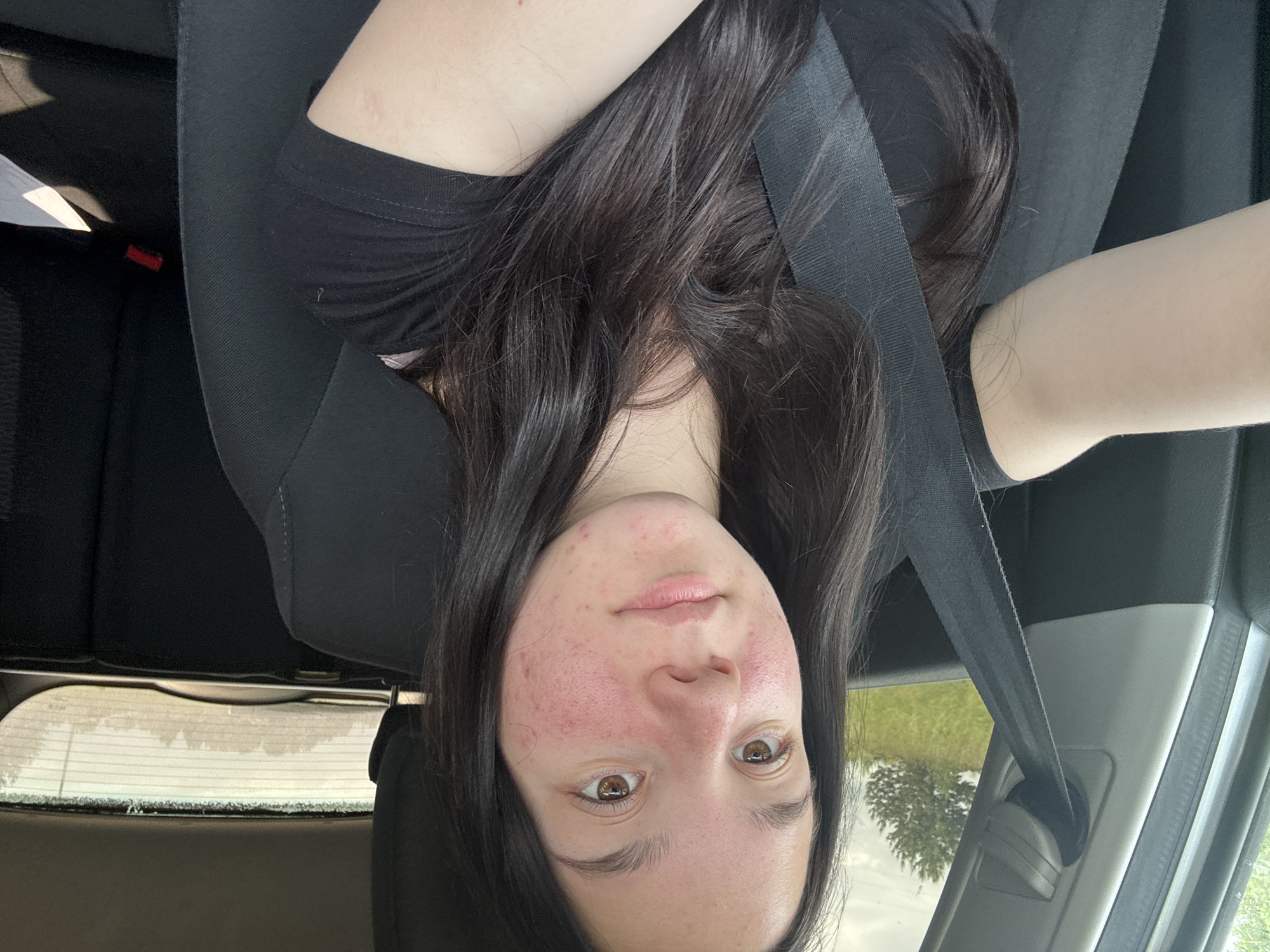 A young woman with dark hair, wearing a black t-shirt, sits in the driver's seat of a car. She is looking directly at the camera, with her left arm extended and resting on the armrest. A black seatbelt is visible across her chest. The background shows the interior of the car, including the passenger seat and headrests, as well as a blurry view of trees and grass through the car windows. The scene appears to be in the city of Nozhovka, Russia.