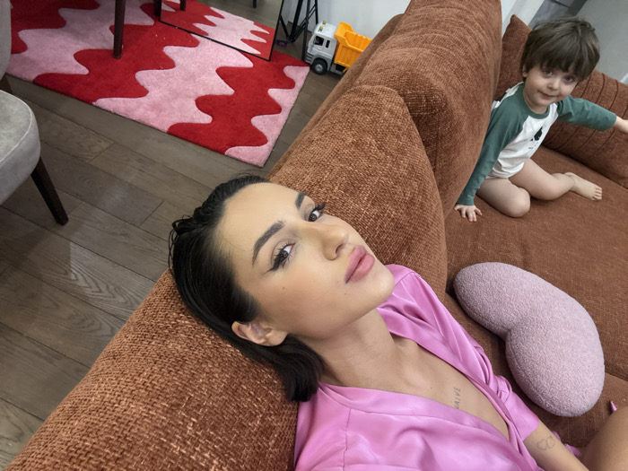 The image displays an intimate indoor scene featuring a woman and a young child relaxing on a brown couch. In the foreground, a woman with dark hair, styled away from her face, lies partially on her side on the plush, textured brown couch. She is looking directly at the camera with a calm expression, sporting dark, well-defined eyebrows, winged eyeliner, and pink lipstick. She is dressed in a vibrant pink, possibly satin, robe or top, which reveals a tattoo on her chest spelling out "NAIVE." A smaller, less distinct tattoo is also visible on her left forearm.
To the right and slightly further back on the same couch, a young child with light brown hair is seated. The child wears a green and white long-sleeved shirt and light-colored bottoms (possibly a diaper or shorts), with bare legs and feet. The child is also looking towards the camera, extending one arm to the side and sporting a slight smile.
A light pink, irregularly shaped (like a bean or kidney) textured pillow rests on the couch next to the woman. The floor beneath the couch is composed of light wooden floorboards. On the floor, a striking red and light pink rug with a bold, wavy pattern is visible, adding a pop of color to the room. Part of a grey upholstered chair with dark wooden legs can be seen on the left side of the image. In the background, near the child, a yellow and grey toy truck is identifiable, suggesting a playful environment. A reflection of the patterned rug and some other elements is visible in what appears to be a mirror or reflective surface in the upper left corner of the scene. The overall ambiance is relaxed and casual, indicative of a home setting, likely a living room. The lighting appears soft and even, suggesting either natural diffused light or indoor lighting.