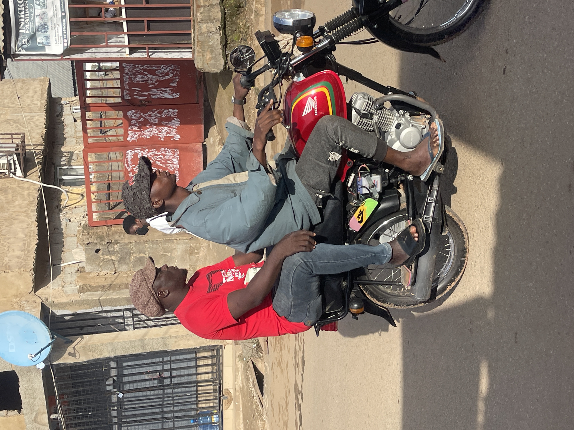 Two men are depicted on a red Honda motorcycle, parked on a paved road in what appears to be an urban or semi-urban setting in Abuja, Nigeria, under bright daylight. The image is taken from a slightly elevated perspective, looking down on the scene.
The driver, wearing a light blue long-sleeved shirt or jacket, grey sweatpants, and blue flip-flops, is positioned in front, his left hand on the handlebar. He wears a dark, textured cap and is looking upwards. The passenger, seated behind him, wears a red short-sleeved t-shirt, blue jeans, and black flip-flops, topped with a brown flat cap. His arms are resting on his lap, and his gaze is directed upwards and slightly away. A third person's head, wearing a white shirt, is partially visible behind the driver.
The motorcycle itself is red with yellow and green stripes on the fuel tank, featuring the "HONDA" logo prominently. The engine, exhaust, and wheels are visible, and the motorcycle appears to be stationary, possibly awaiting something or someone. The sun casts strong shadows of the motorcycle and people onto the road surface, indicating a clear, sunny day, likely around midday or early afternoon.
In the background, there are several buildings made of concrete or stone, suggesting typical local architecture. A prominent feature is a red metal gate with white, graffiti-like markings. Above this gate, a white sign is visible with black text that reads "INIKKO FASHION" and a phone number "+61 69958038", along with other smaller, less discernible text likely detailing services like "Corporate wears" and "Casual." To the far left, a light blue satellite dish is mounted on a building. The overall scene suggests a common street view in a bustling residential or commercial area.