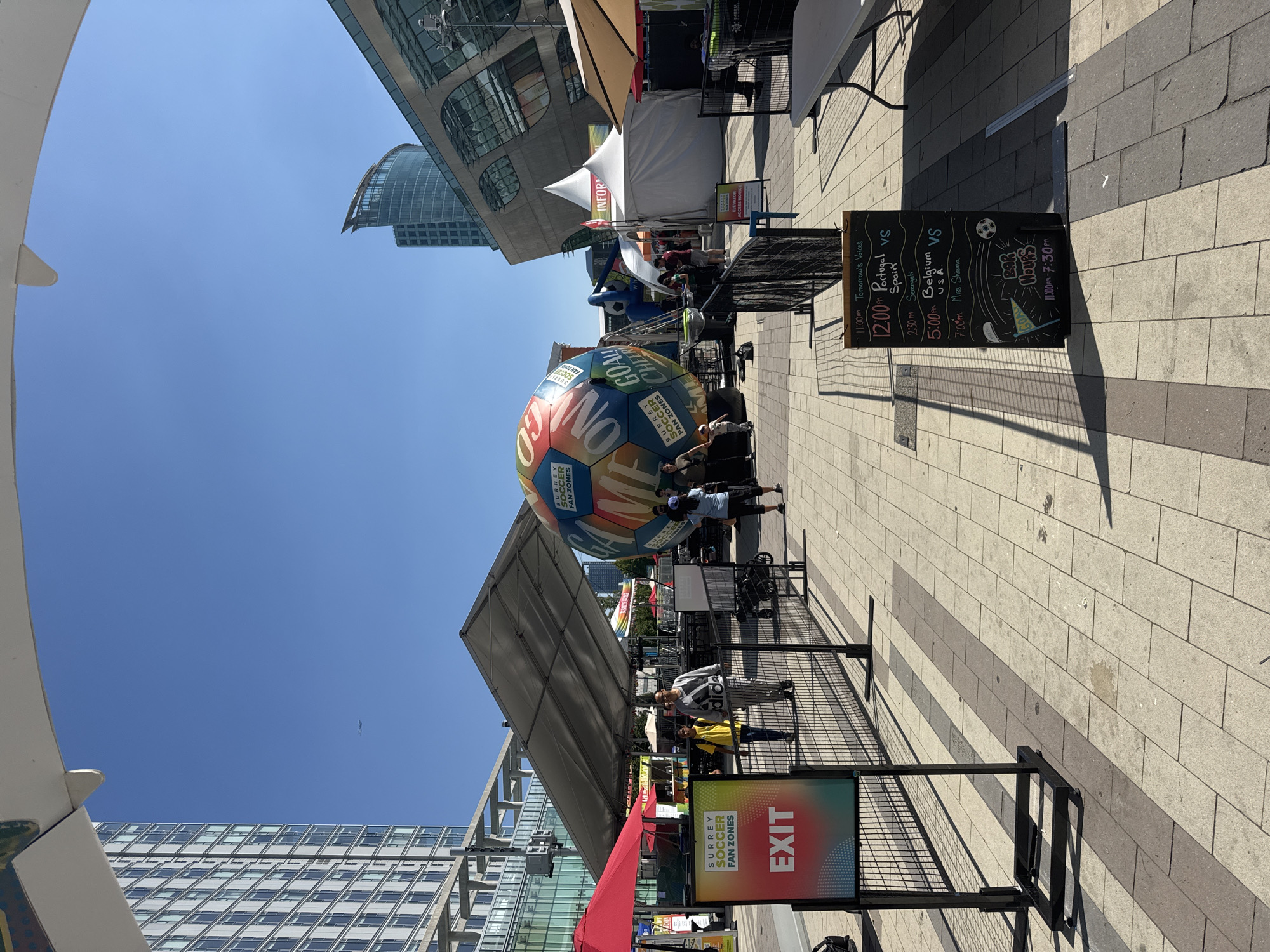 In Surrey, Canada, a vibrant outdoor fan zone is set up on a sunny day under a clear blue sky. The scene is framed by a large white architectural overhang. In the background, a tall, modern glass skyscraper stands on the left, and another distinctive, curved glass building is visible on the right, suggesting an urban environment.
The central focus is a massive, inflatable soccer ball adorned with colorful panels bearing text like "GOAL," "CHEER," and "SURREY SOCCER FAN ZONES." The ball is positioned near a large, open-sided tent structure with a gray roof, under which tables and chairs are set up, indicating a space for events or gatherings. Several white tents, possibly for vendors or information booths, are also present.
People are scattered throughout the area. Two individuals are walking towards the viewer, one in a yellow shirt and the other in a gray tracksuit with "adidas" branding. Further back, a group of people are gathered near the tents. A child is seen near the inflatable ball, and other adults are standing or walking around.
In the foreground, a black metal fence separates the viewer from the fan zone. A sign with "SURREY SOCCER FAN ZONES" and "EXIT" in bright, gradient colors is prominently displayed on the left. A blackboard easel stands on the right, displaying a schedule of events including soccer match times (e.g., "12:00pm Portugal VS Spain," "5:00pm Belgium VS USA") and "BAR HOURS" from 11:00 AM to 7:30 PM. The ground is paved with interlocking rectangular tiles in shades of gray and beige. The overall atmosphere suggests a lively event, likely related to a soccer tournament.