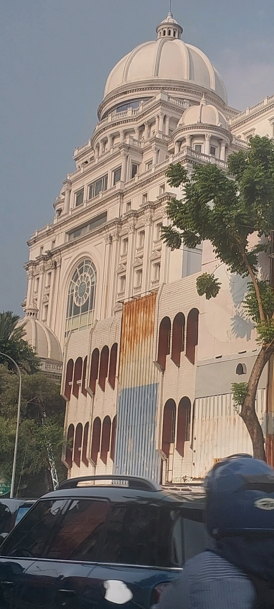 The image shows a close-up view of the building of the PT Telkom building in Surabaya, Indonesia. The building is a tall, white, classical-style building with a prominent dome and ornate architectural details. In the foreground, there are cars and a person on a motorcycle, suggesting a street scene in a busy urban environment. The sky is clear and light blue. The building's facade features arched windows and decorative columns. The overall impression is of an impressive and historic structure set against a backdrop of everyday city life.
