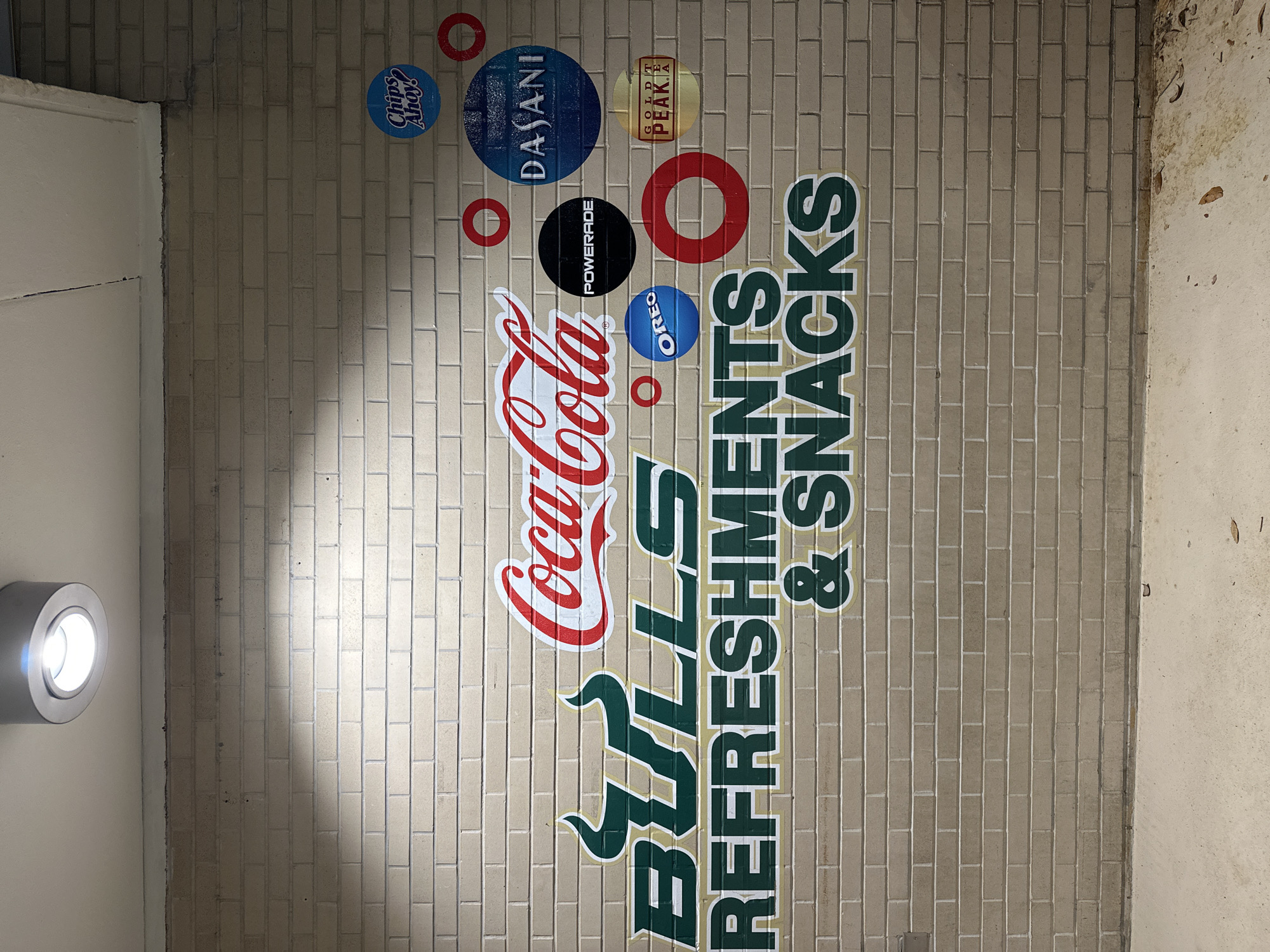 The image depicts an advertisement for Bulls Refreshments & Snacks, prominently featuring the Coca-Cola logo, along with other brand logos such as Dasani, Powerade, Oreo, Gold Peak Tea, and Chips Ahoy! The sign is affixed to a brick wall, likely within a retail or concession area. The location is identified as being within the city of Tampa, United States of America. The advertisement consists of a series of circular logos and brand names arranged vertically. The Bulls logo is positioned below the Coca-Cola logo, followed by the words "Refreshments & Snacks" stacked vertically. A circular light fixture is visible on the wall next to the brick wall, and there is a plain white surface to the left of the sign. The area to the right of the sign appears to be a decaying wall.