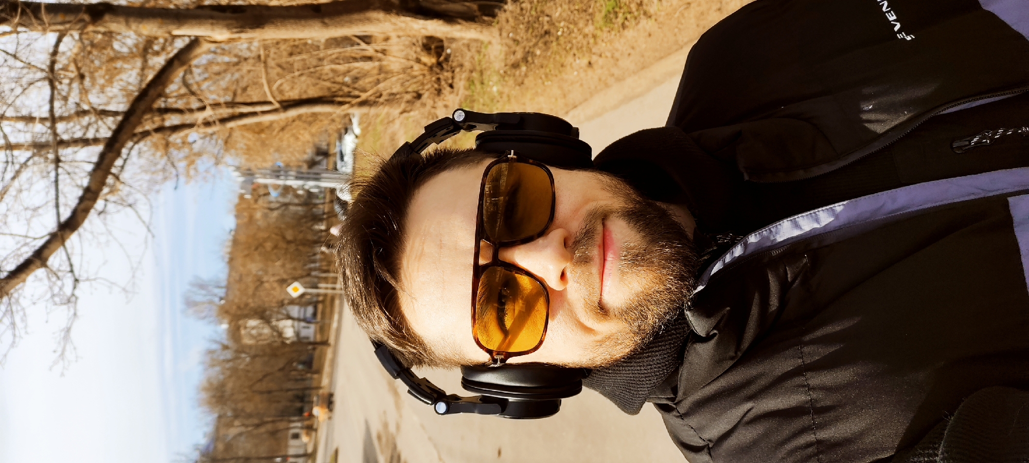 A close-up, slightly angled headshot of a man with a beard wearing sunglasses and headphones. He is positioned outdoors on what appears to be a paved path. Bare trees are visible in the background, along with buildings and street signs. The lighting suggests it is daytime. The location is Fryazino, Russia, as indicated by a street sign. He's wearing a black jacket, partially unzipped.