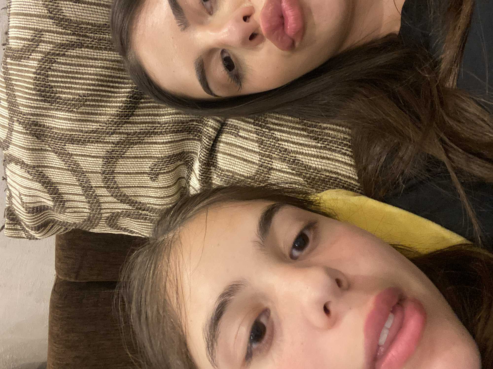 Two women are lying down next to each other. The woman on top is resting her head on a striped pillow. The other woman is lying on a brown sofa and wearing a yellow garment. The location is not discernable from the given image.