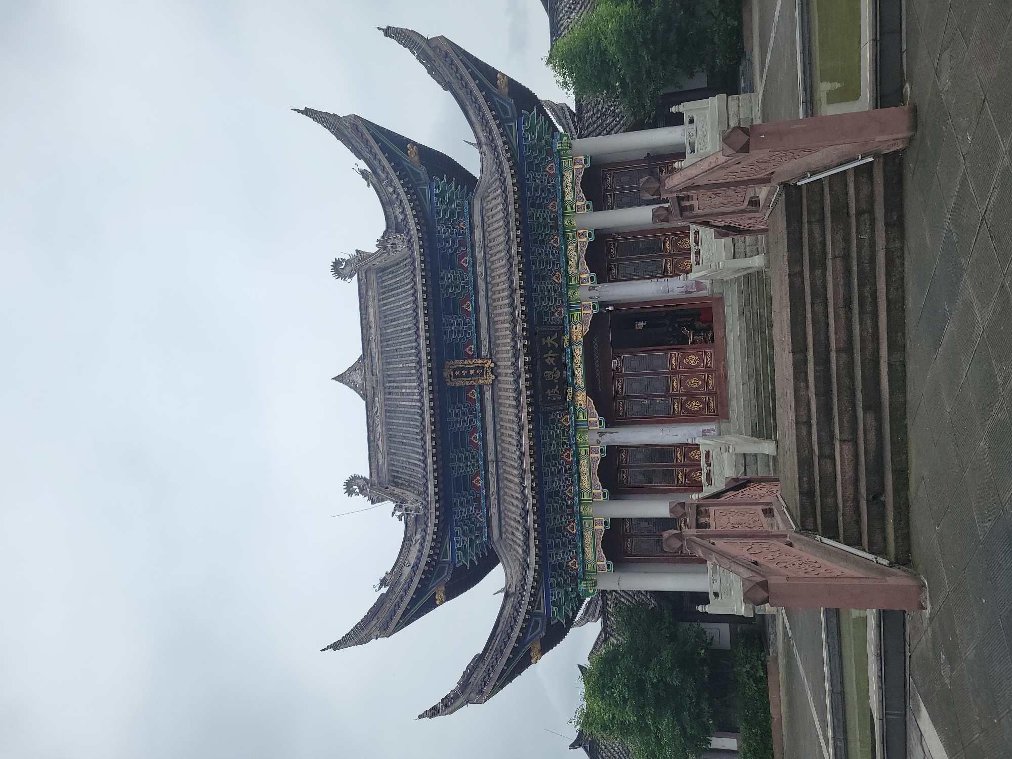 The image shows the Wenfeng Pagoda in Hechuan, China. The pagoda has a traditional Chinese architectural style, with curved eaves and intricate decorations. The building is tall and imposing, with multiple stories. It has gray tiles and blue and green painted accents. It is approached by a set of stone stairs with red railings. The sky is overcast. Trees are visible adjacent to the pagoda.