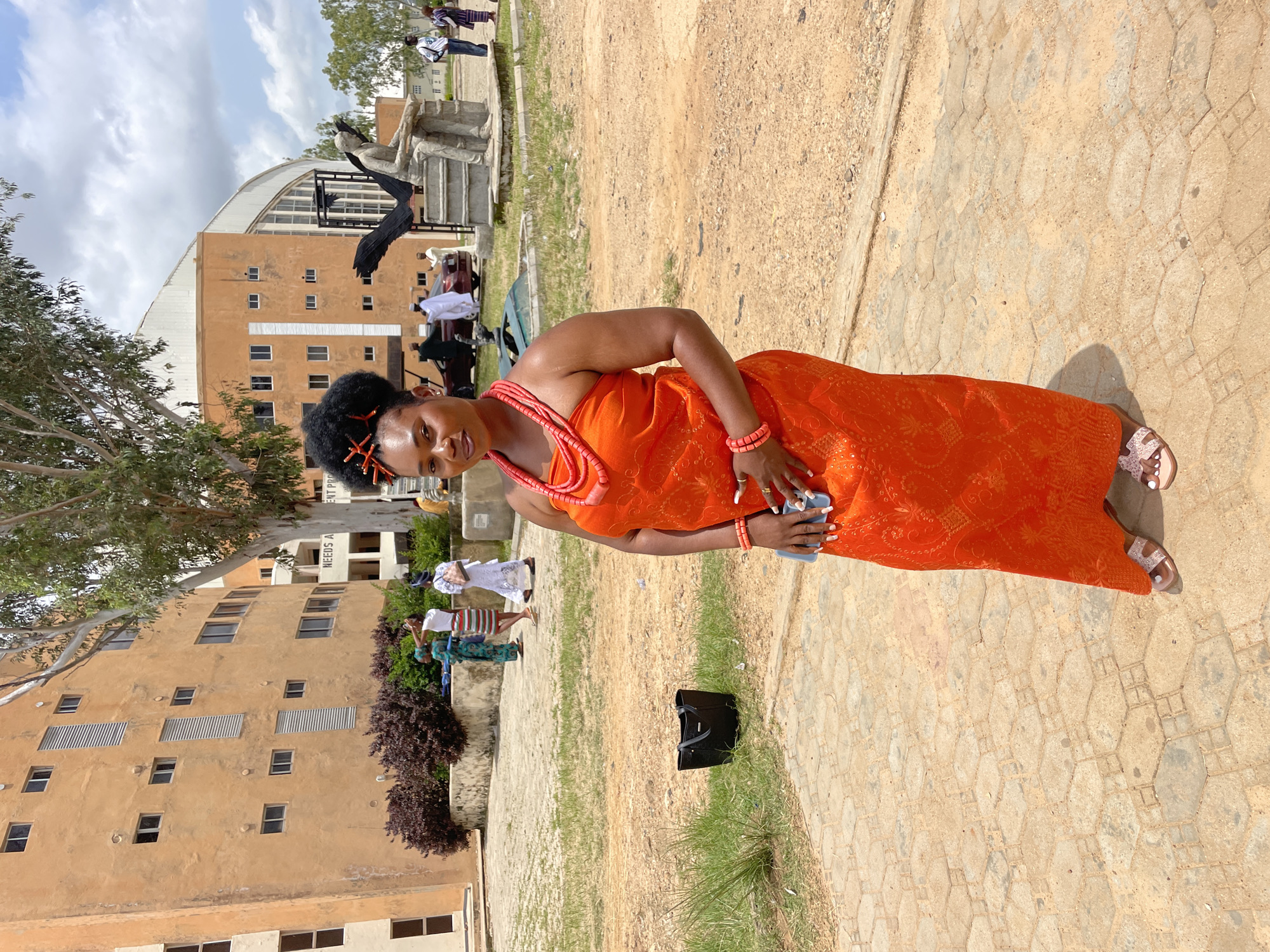A woman stands in the foreground, facing forward and slightly to the left. She is wearing a vibrant orange strapless dress with intricate patterns, a matching beaded necklace, and beaded bracelets on her wrists. Her hair is styled in an updo with red decorative pieces. She holds a light blue mobile phone in her right hand. Her feet are adorned with pink sandals. Behind her, a black handbag rests on the ground.
In the mid-ground, several other people are visible. Two women in traditional attire, one in a green and blue patterned dress and another in a white dress with a striped skirt, are walking away from the camera. Further back, a man in a white garment is seen near a car. To the right, a statue of a seated man holding what appears to be a bird and a larger bird sculpture are situated in front of a curved, orange-colored building with arched windows.
The environment appears to be an outdoor public space, possibly a plaza or courtyard, with a paved ground composed of hexagonal tiles in the foreground and sandy earth in the background. A large tree with sparse foliage stands behind the woman. In the distance, additional buildings are visible under a cloudy sky. The overall scene suggests an event or gathering taking place in an urban setting. The curved orange building in the background, with its distinctive architecture, is characteristic of the National Museum Jos, a landmark in the city of Jos, Nigeria.
