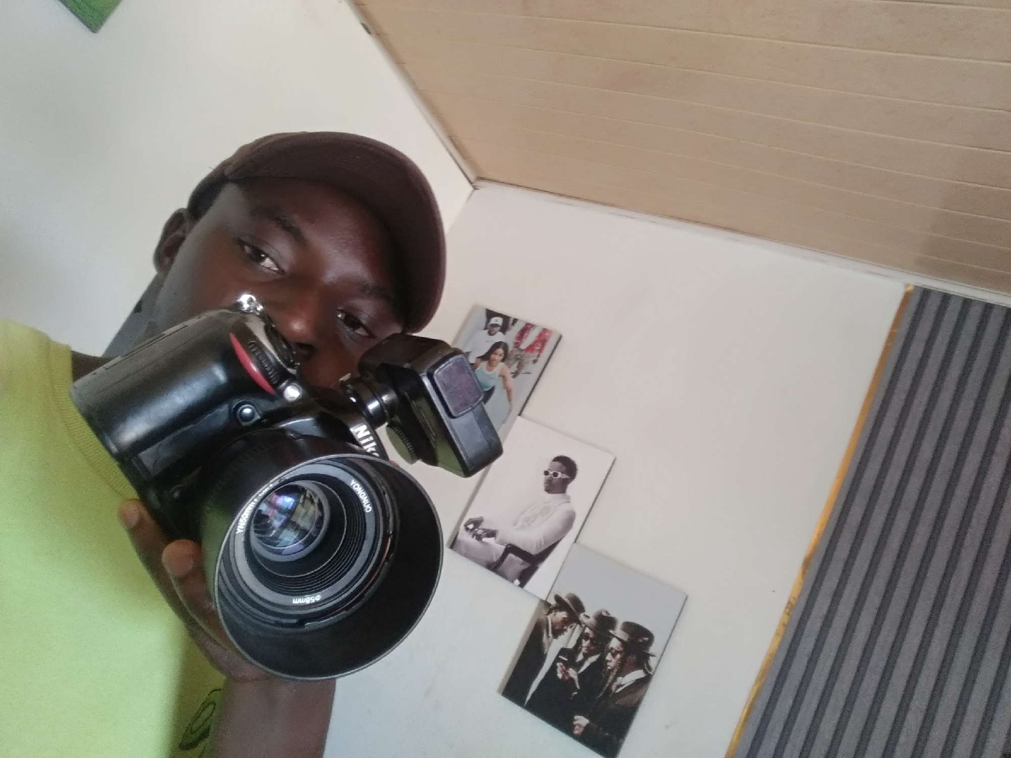A man with dark skin and a brown cap is captured in a close-up, holding a black Nikon DSLR camera with a Yongnuo lens and an attached external flash unit. He is wearing a light green t-shirt and is looking directly at the camera, suggesting he is taking a selfie or recording. The camera's lens clearly displays "YONGNUO" and "Φ58mm" text.
The indoor setting features a light-colored wall in the background, adorned with three framed images. The top picture shows a woman in a light-colored top, the middle one depicts a man in white attire and sunglasses, and the bottom image features three men in hats and dark clothing, resembling Hasidic Jews. Above the wall, a light brown paneled ceiling is visible, and to the far right, a vertical striped pattern in grey and white, possibly a wallpaper or curtain, adds texture to the background.