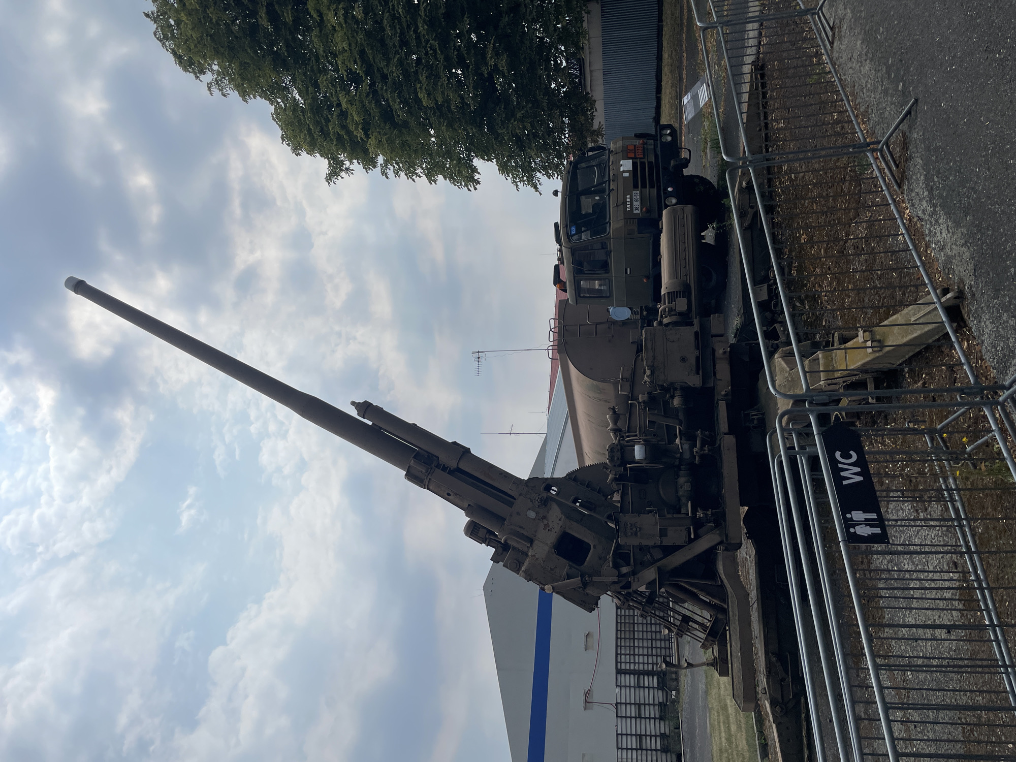 In the city of Unknown, Unknown, an outdoor exhibition features a large, weathered military artillery piece positioned on a mobile platform. The artillery piece has a long barrel angled upwards, and is connected to a truck with a large tank. The truck is olive drab and bears markings including "Tatra 383 60-59" and other Cyrillic script. The scene is set against a backdrop of a cloudy sky and a large, modern building with blue trim. A chain-link fence encloses the exhibit, and a sign indicating "WC" with pictograms for men and women is visible on the fence. A mature tree with dense green foliage stands to the right of the scene, and a grey, corrugated metal fence runs along the background. The ground is paved and covered with gravel and scattered debris.