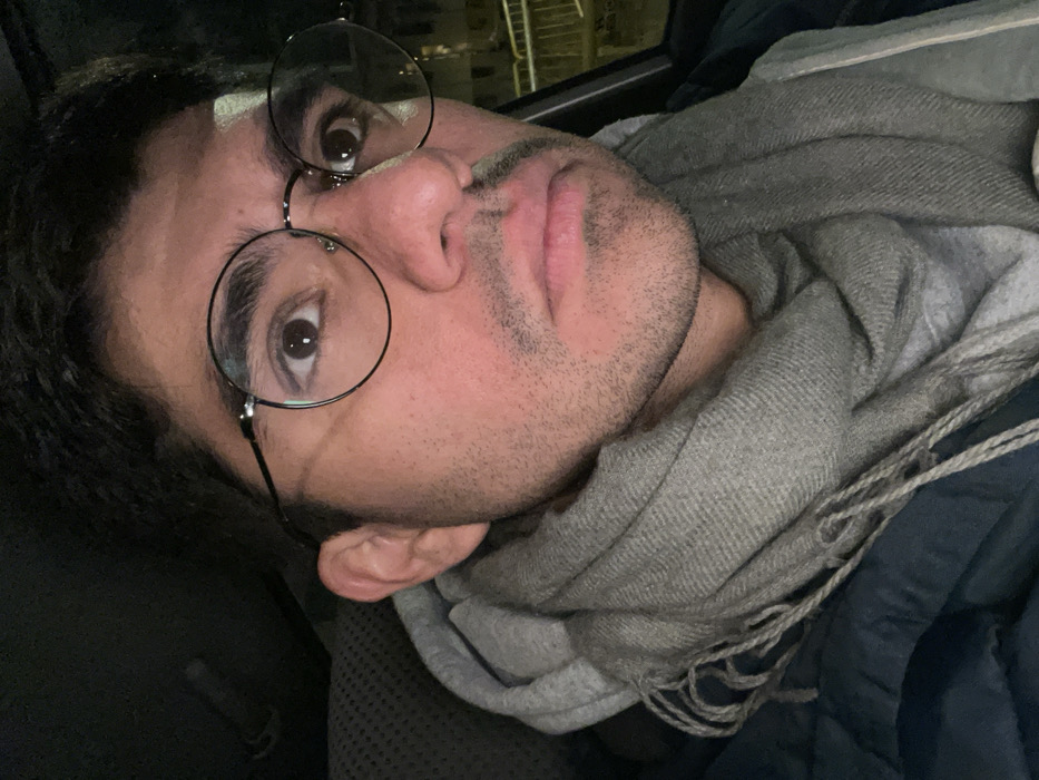A man with dark hair and stubble wears round glasses and a gray scarf. He is lying down, possibly in a car, with a dark background visible through the window. A blue jacket or blanket is also present. The man is looking slightly upward toward the camera, his gaze visible through the lenses of his glasses.