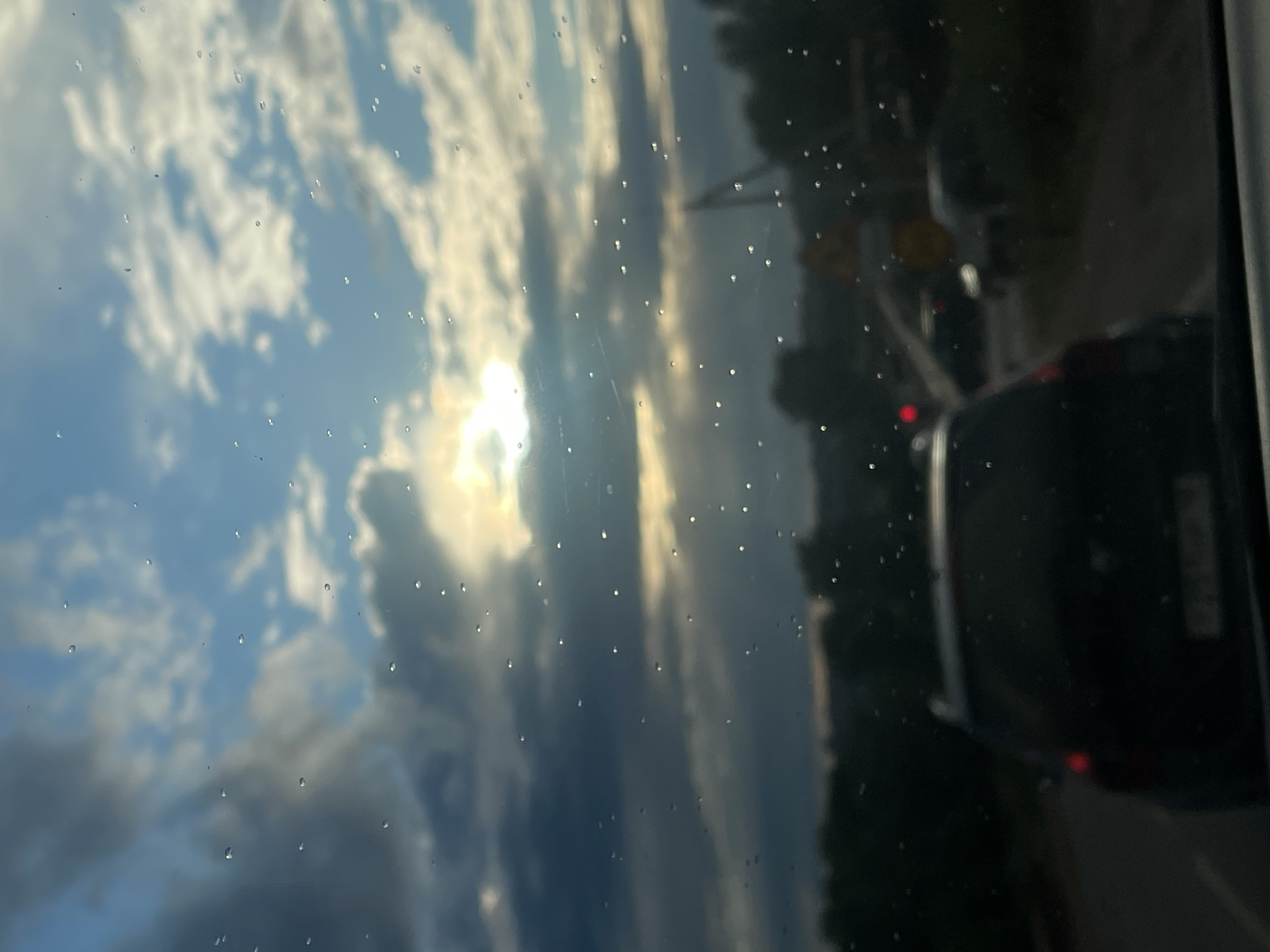 The image depicts a view through a wet window, likely from inside a vehicle. The subjects are a cloudy sky with the sun partially obscured by clouds, and a reflection of traffic on a road. We see the back of a black SUV and other vehicles in the distance. The scene is set on a road, indicated by the traffic and a yellow traffic sign visible on the right. There are numerous water droplets on the window, suggesting it is raining or has recently rained. The lighting indicates it is daytime, likely late afternoon or early evening due to the warm, diffused sunlight. The overall mood is somewhat somber due to the weather, but the sun's rays offer a hint of brightness. No discernible text is visible. The location is identified as Gavrilovka, Russia.