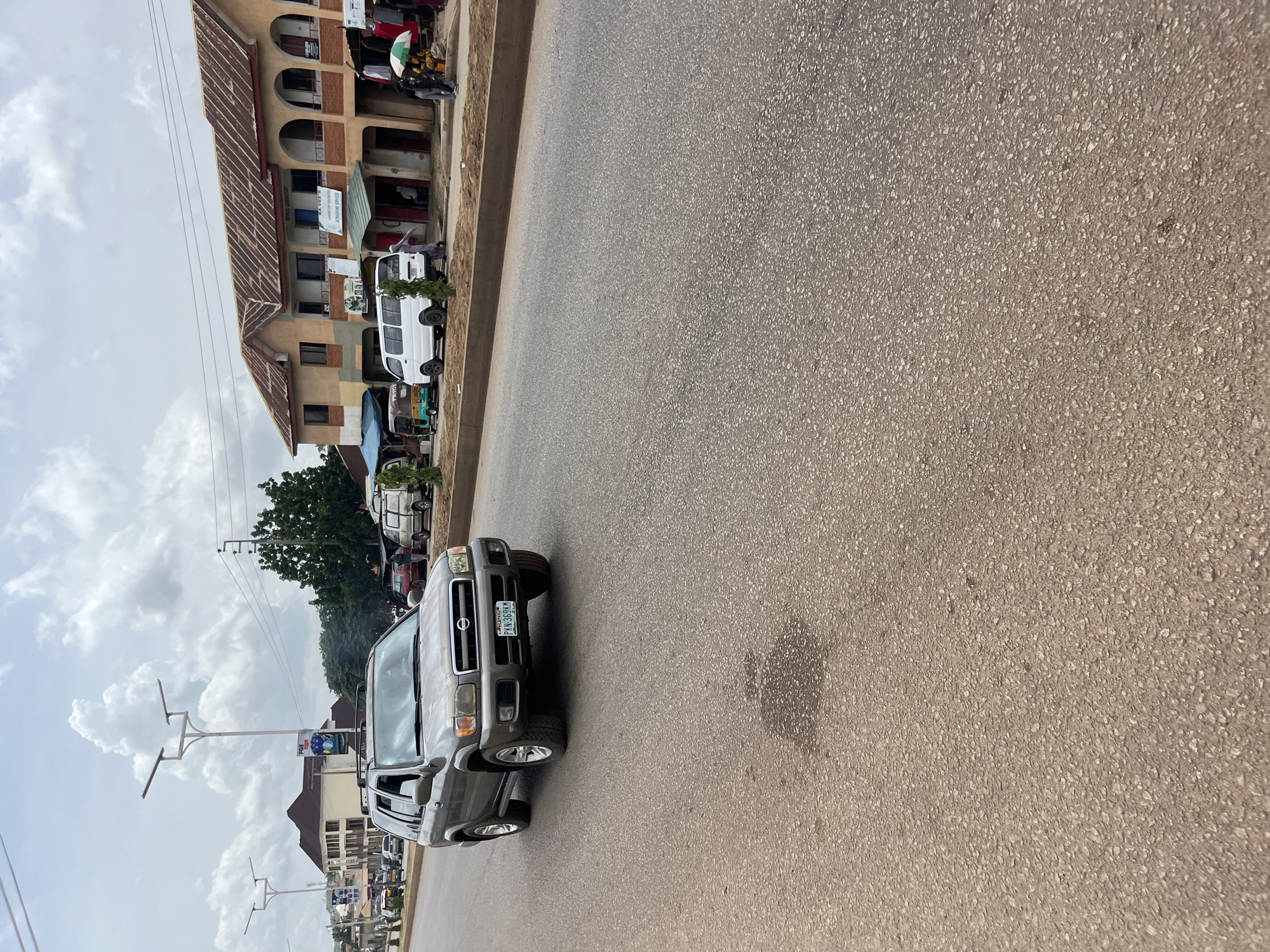 In Bukuru, Nigeria, on a street with asphalt pavement and a dirt shoulder, a grey Nissan Pathfinder SUV is driving on the road. The SUV, with license plate "PKN 369KM," is in the left lane, moving towards the viewer. On the right side of the road, a two-story building with a rusted, corrugated metal roof houses various businesses, including "RA-VEES LABORATORY IMAGING" and "ULTRASONIC SERVICES." A white van, a yellow and green tricycle, and pedestrians are also present on the right side of the road. The sky is overcast with clouds. Streetlights and overhead wires are visible in the background.