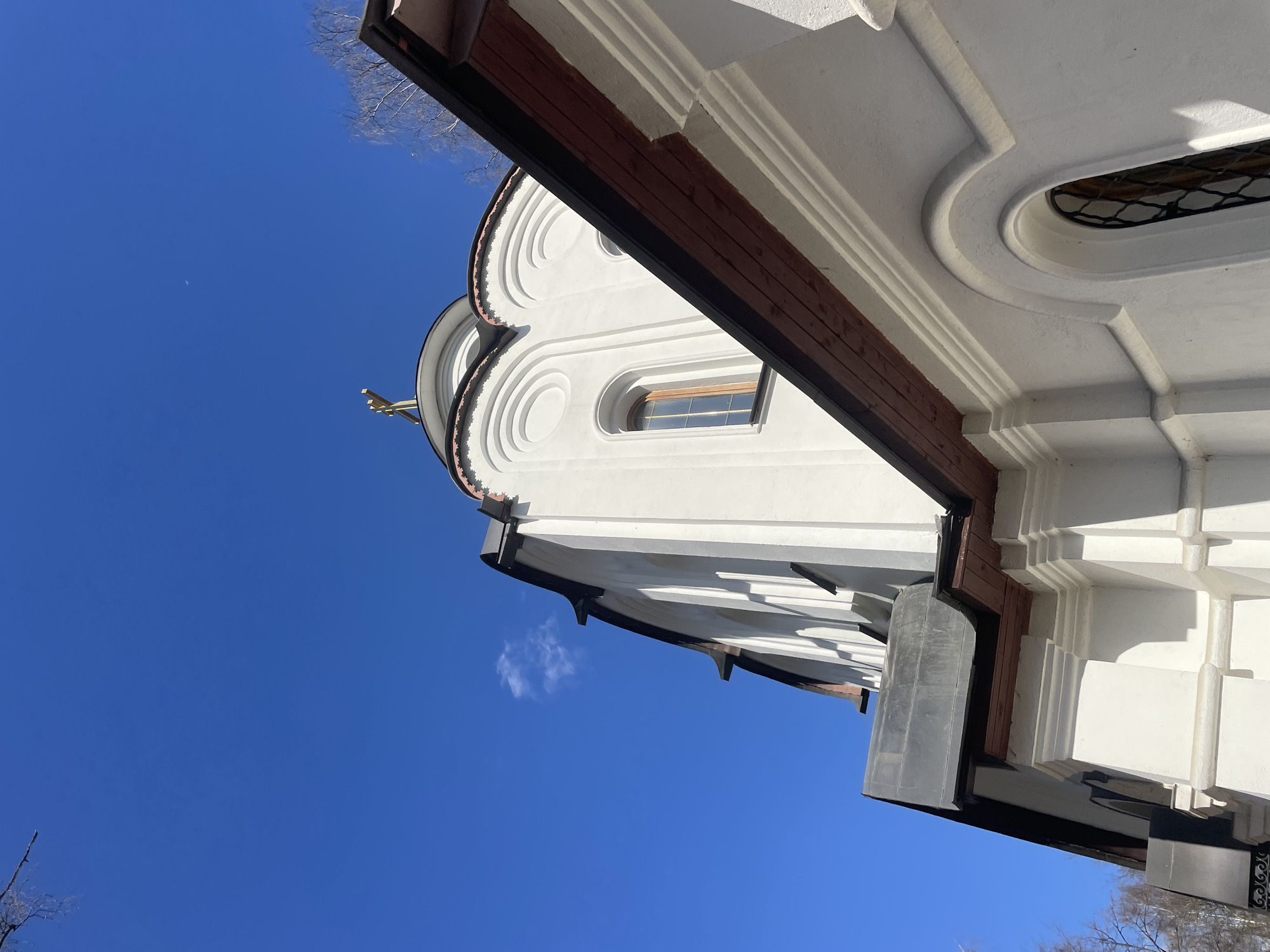 Here is a description of the image:
The image shows the exterior of a white building, likely a church or religious structure, viewed from a low angle looking upwards. The building features rounded architectural details, decorative molding, and a cross on its roof. The building appears to be constructed of white-painted stone or plaster with dark brown trim along the edges. There are window openings visible, some rectangular and one oval. The background is a clear blue sky with a few bare tree branches visible at the edges of the frame. The composition is angled, giving a dynamic perspective.
Based on the architectural style, it is possible this building is located in Moscow, Russia. However, without additional visual clues or context, this is a preliminary guess.