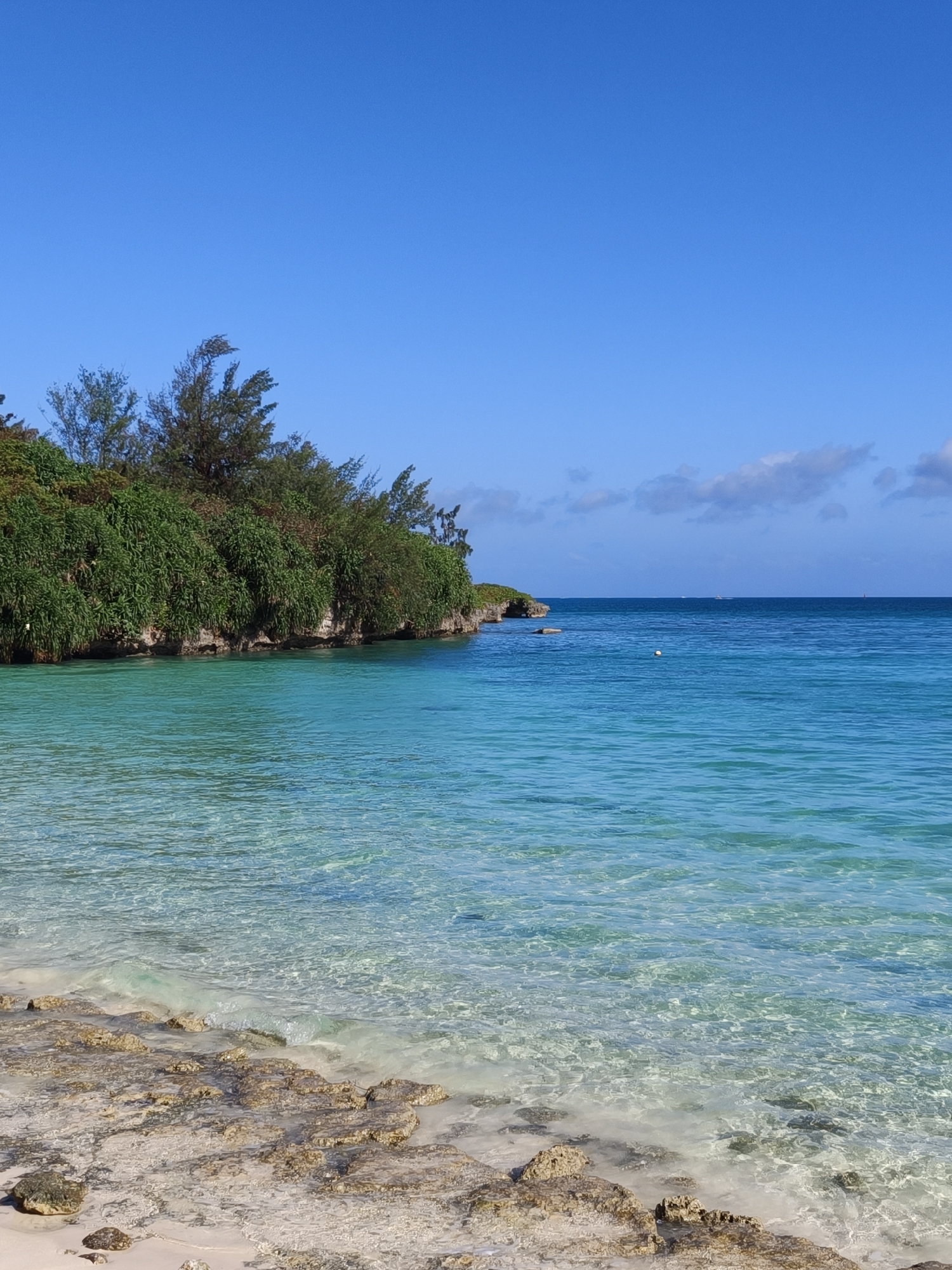 The image shows a scenic view of the ocean and coastline. In the foreground, a sandy beach with scattered rocks meets the clear, turquoise water. The water is shallow near the shore, allowing the seabed to be visible. Further out, the ocean transitions to a deeper blue, with a few distant boats and a buoy visible on the surface. The coastline is characterized by a rocky cliff covered in lush green vegetation and trees. The sky is a clear, vibrant blue with a few scattered clouds. This scene is reminiscent of Yonaha Maehama Beach in Miyakojima, Japan, known for its stunning white sand and crystal-clear waters. There are no people or animals visible in the image.