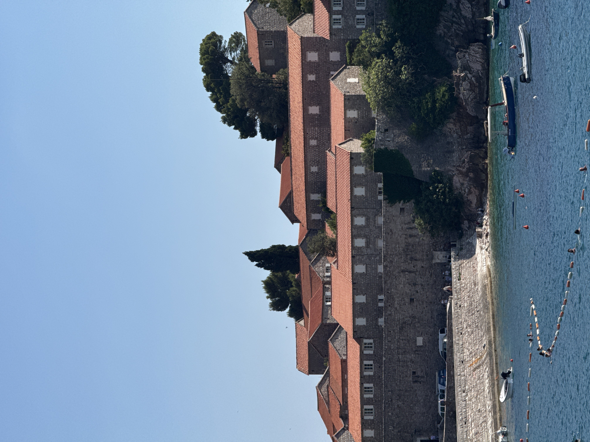 This image shows a scenic view of Sveti Stefan, Montenegro, on a clear, sunny day. The scene is dominated by a row of stone buildings with distinctive red-tiled roofs, clustered together on a rocky outcrop. These buildings appear to be historic, possibly part of a resort or old village. Lush green trees are interspersed among the structures, adding a natural element to the architectural landscape.
Below the buildings, the turquoise waters of the Adriatic Sea lap at a stone-paved shoreline. Several boats are visible in the water, including a white motorboat and a darker vessel with an awning. Mooring buoys are present, indicating a busy marine environment. Some people are seen near the shore, with a few individuals swimming or enjoying the water.
The time of day appears to be daytime, with bright sunlight casting shadows and illuminating the scene. The overall atmosphere suggests a relaxed, vacation-like setting, characteristic of a popular coastal destination. The location cues, such as the unique architecture and the clear blue sea, strongly suggest Sveti Stefan. There is no visible text in the image.