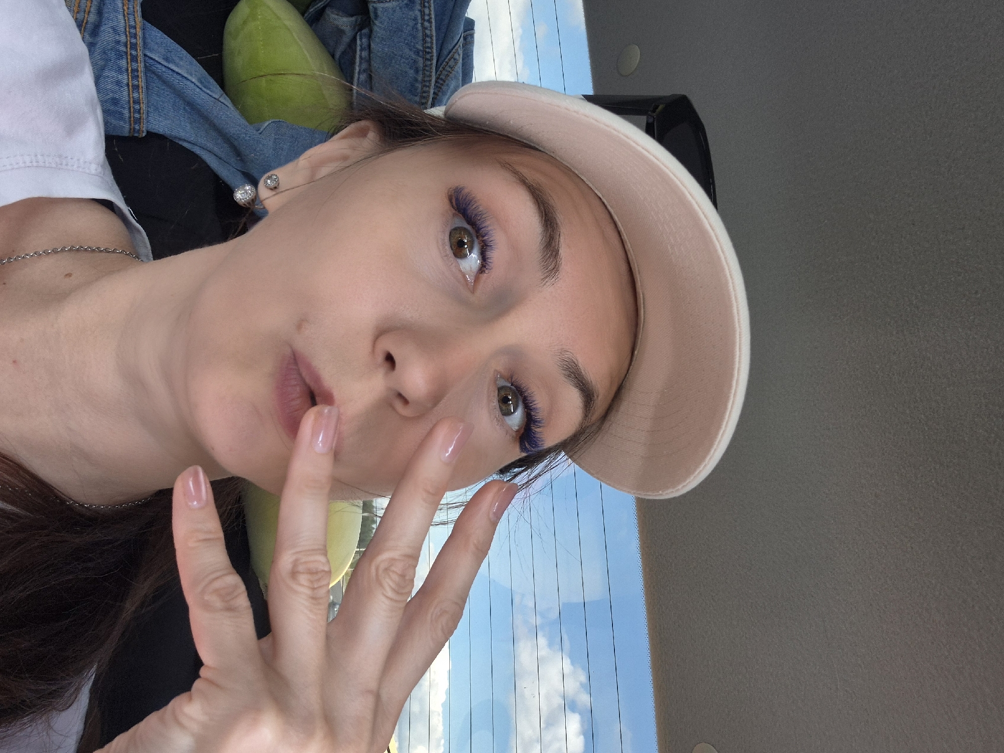 In Novoalekseevskoe, Russia, a young woman with purple eyelashes and a pink baseball cap is taking a selfie inside a vehicle. She is making a kissing face and holding up her hand, which has clear nail polish. A blue denim jacket and a green object are visible behind her. The background shows the sky with clouds through the car's rear window.