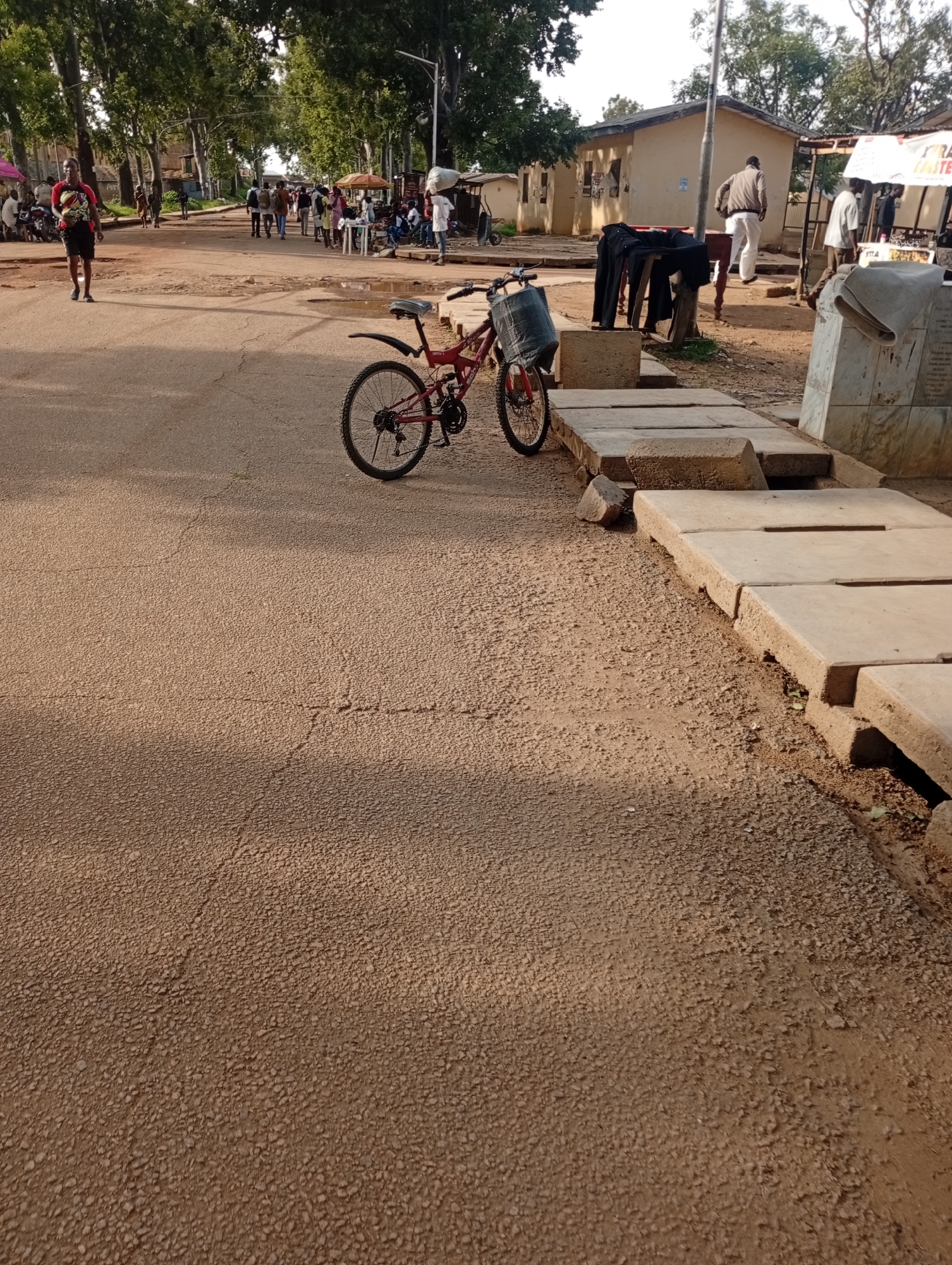 The image depicts a dusty, unpaved street scene in what appears to be Rafin Par, Nigeria. A red bicycle with a basket is parked on the right side of the road. Several people are walking in the background, and some are gathered near stalls or shops. Buildings with light-colored walls line the street. A large tree with dense foliage stands on the left side of the image. The ground is covered in a granular, reddish-brown material. There are also some concrete structures and a monument on the right side of the road. The lighting suggests it is daytime, with shadows cast on the ground.