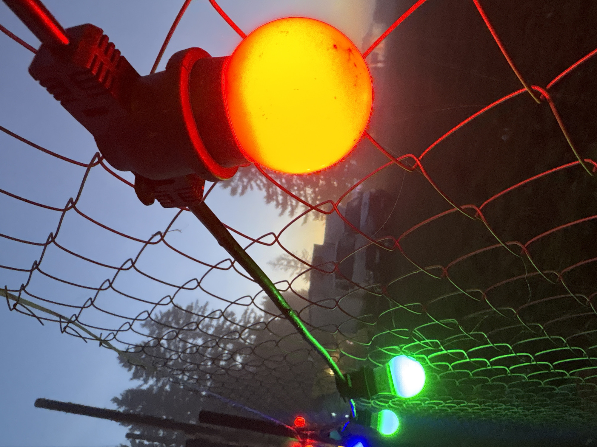 The image depicts a close-up of a chain-link fence in Polevaya, Russia, during twilight. String lights with red, yellow, and green bulbs are attached to the fence, illuminating the scene. The fence is draped with the lights, with one prominent yellow bulb in the foreground casting a warm glow. In the background, the silhouettes of evergreen trees are visible against a hazy, pale blue sky, suggesting a misty or foggy environment. A dark vehicle, possibly a van, is faintly discernible behind the fence. The overall atmosphere is serene and slightly ethereal due to the low light conditions and the colorful illumination from the string lights.