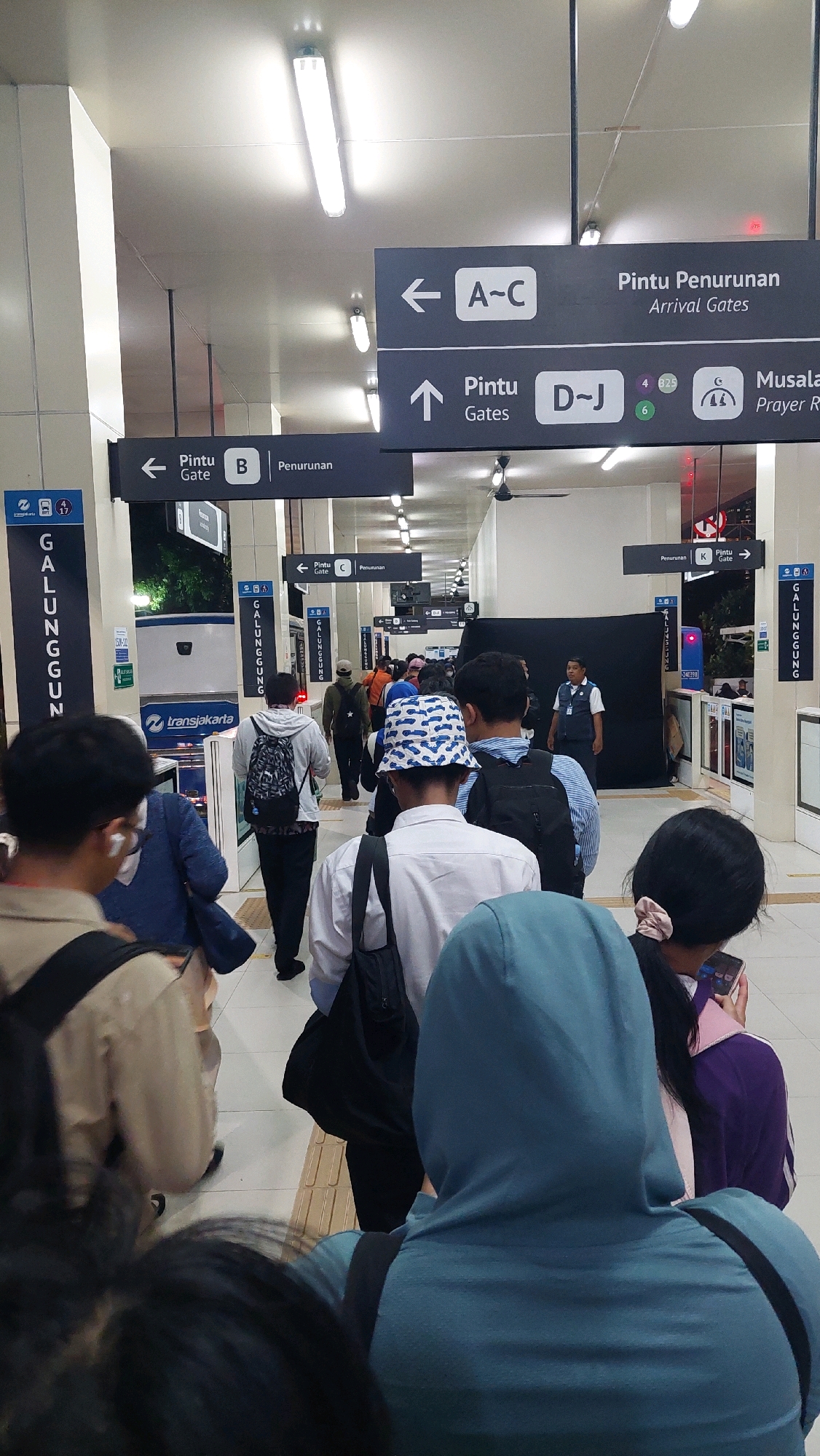The image depicts an indoor scene, likely a transportation hub such as a train station or bus terminal, in South Jakarta, Indonesia. The perspective is from behind a group of people standing in a queue. Overhead, there are signs in Indonesian and English indicating directions to different gates and areas, with some signs pointing to "Pintu Penurunan" (Arrival Gates) and others to specific gates like "A-C," "B," "C," and "D-J." Vertical signs with "GALUNGGUNG" are visible on the left and right sides of the image, which is a street and area in South Jakarta. People are dressed casually, many wearing backpacks, suggesting they are commuters. Some individuals are looking at their phones. The environment appears clean and modern, with tiled flooring and fluorescent lighting. The overall atmosphere suggests a busy, functional public space.
