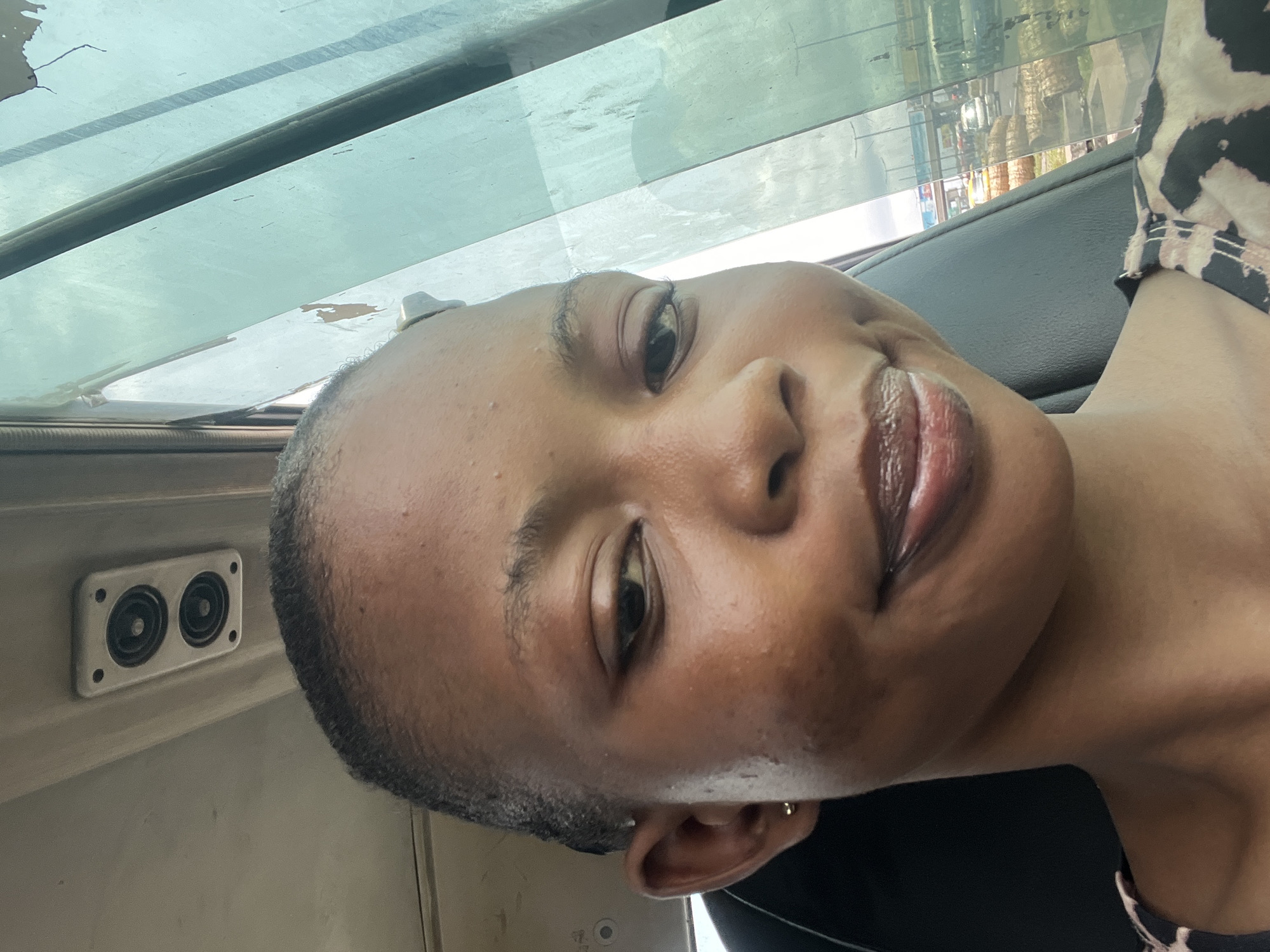 A portrait-style selfie features a young adult with dark skin and very short, dark hair, possibly a buzz cut. The subject looks directly at the camera with a calm, smiling, or smirking expression, showcasing full, glossy lips and a small gold stud earring in their left ear. The image is notably rotated 90 degrees counter-clockwise, with the subject's head tilted to the left.
The setting is inside a vehicle, likely a car or bus, during daytime in Satellite Town, Nigeria. Above and behind the subject's head, a large, clear or lightly tinted window reveals a blurred outdoor scene with green foliage from trees and indistinct buildings or structures, suggesting an urban or suburban environment. The vehicle's interior is visible, with dark grey or black surfaces around the subject's shoulder and neck. To the left, a distinct light-colored panel holds a rectangular unit containing two circular speakers. The subject is wearing a patterned top, possibly an animal print, in black and light brown/cream, visible on the right side of the frame. The natural daylight indicates daytime. No text is visible in the image.