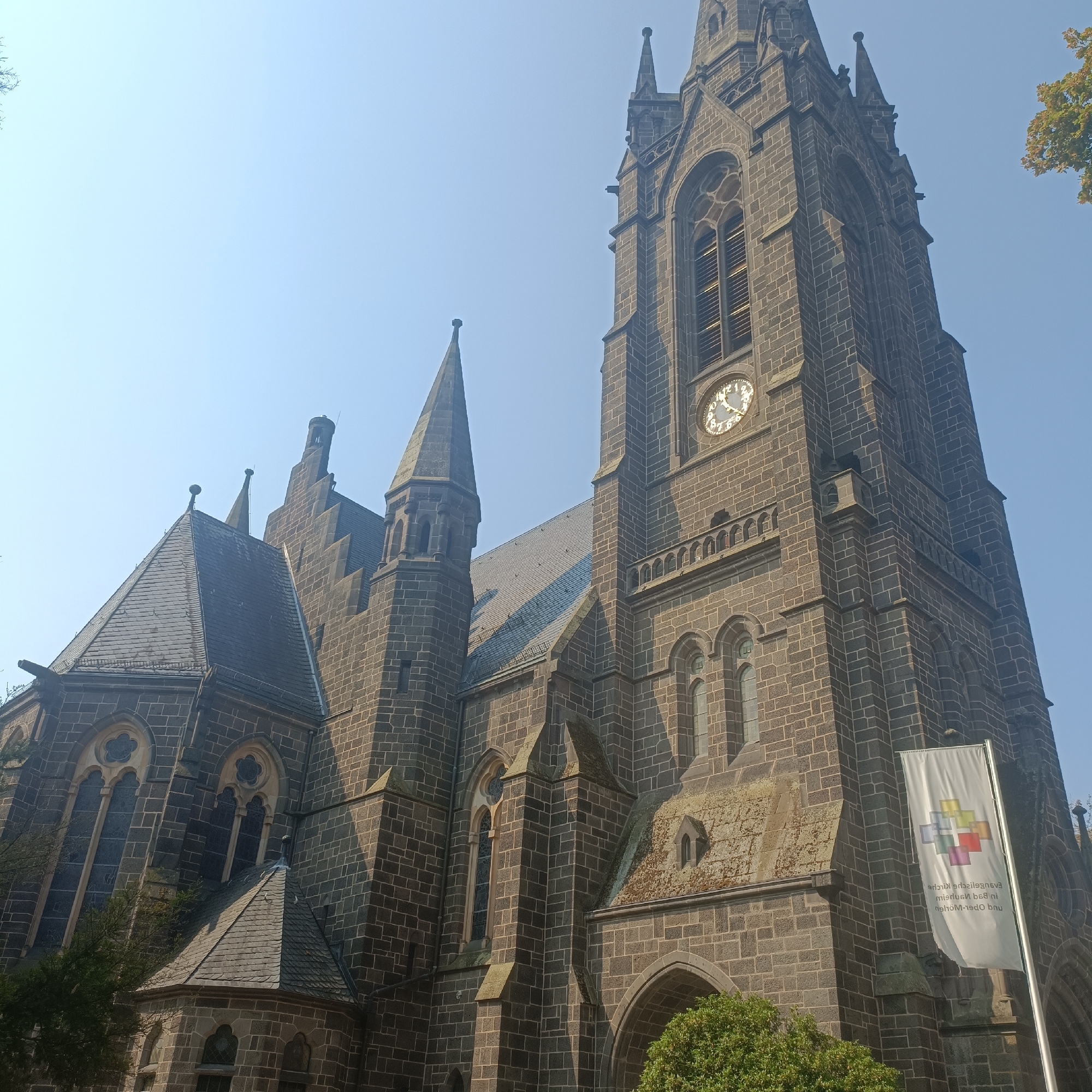 The image shows a low-angle view of the exterior of the Friedenskirche in Bad Nauheim, Germany. The church is constructed from dark stone blocks and features Gothic architectural elements, including pointed arch windows and spires. A prominent clock face is visible on one of the towers. A white banner with a colorful logo and text is displayed on a flagpole to the right of the church entrance. The sky is clear and blue. Green foliage and trees are visible in the foreground and to the right of the church.