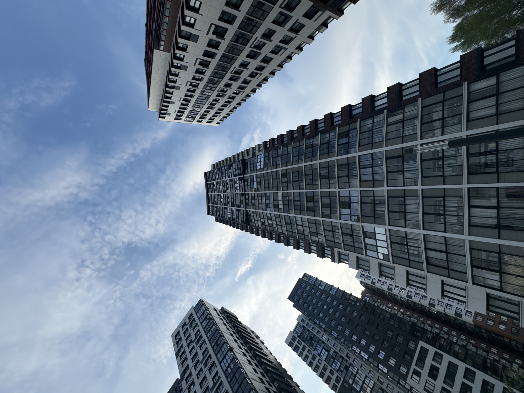 The image is a low-angle shot of several modern high-rise buildings in the city of Odintsovo, Russia. The buildings have a similar style, with a mix of dark and light facades, and large rectangular windows. The sky is partially cloudy, with a mix of blue and white. Some tree leaves are visible at the top right corner.