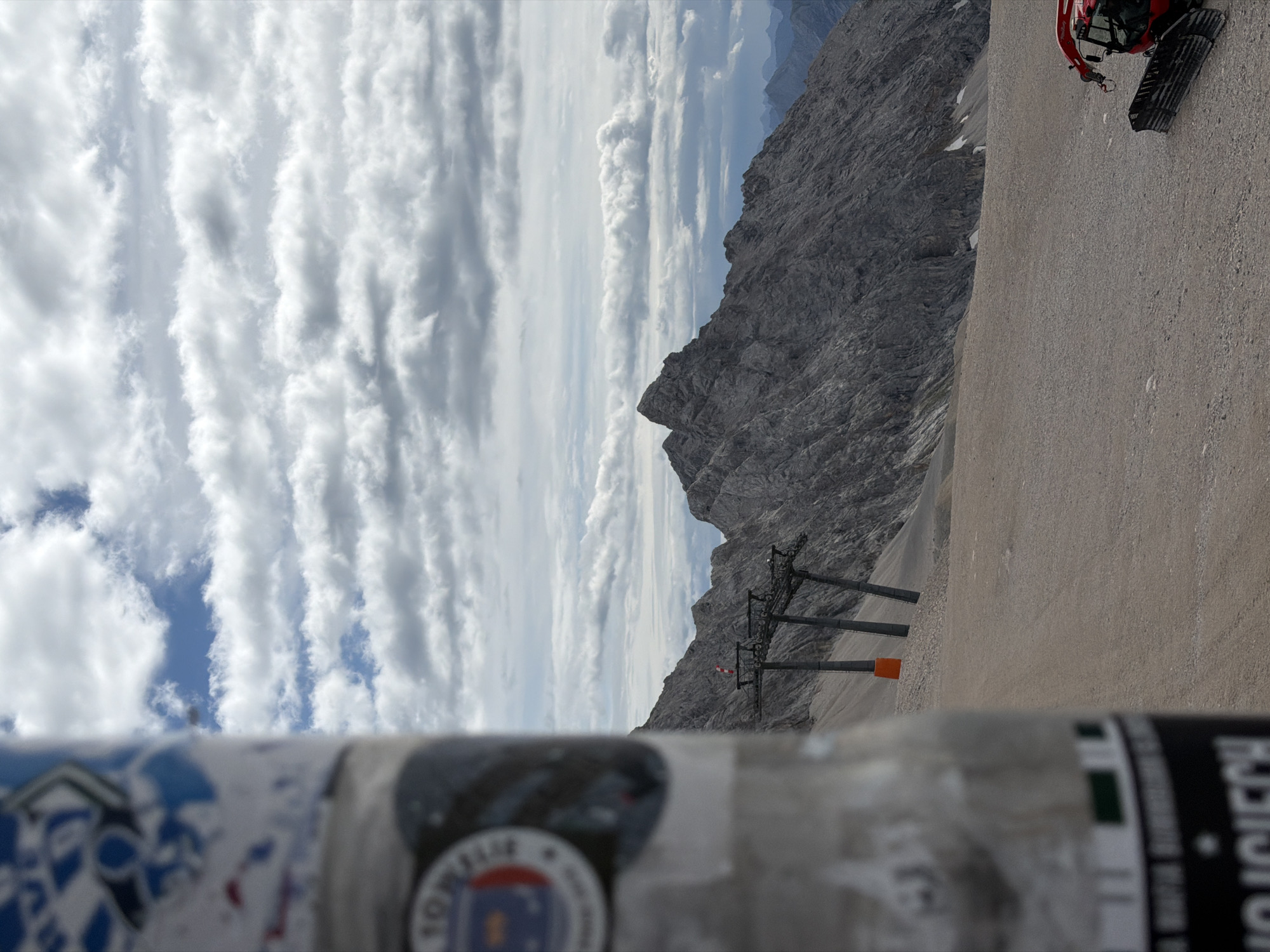 The image depicts a mountain landscape, likely in the Bavarian Alps, near Garmisch-Partenkirchen, Germany, given the context of the photo series from which it is derived. In the foreground, a blurred cylindrical object, possibly a pole or pillar, is covered with stickers. Beyond this, a vast, gravelly expanse stretches towards a rugged, rocky mountain range under a cloudy sky. A ski lift structure with multiple towers is visible in the mid-ground, with orange markers on one of the towers. Further in the distance, more mountain peaks fade into a hazy blue. To the far right, a red snow groomer is parked on the gravelly ground. The scene appears to be a high-altitude location, possibly a ski resort area during the off-season or a scenic viewpoint.