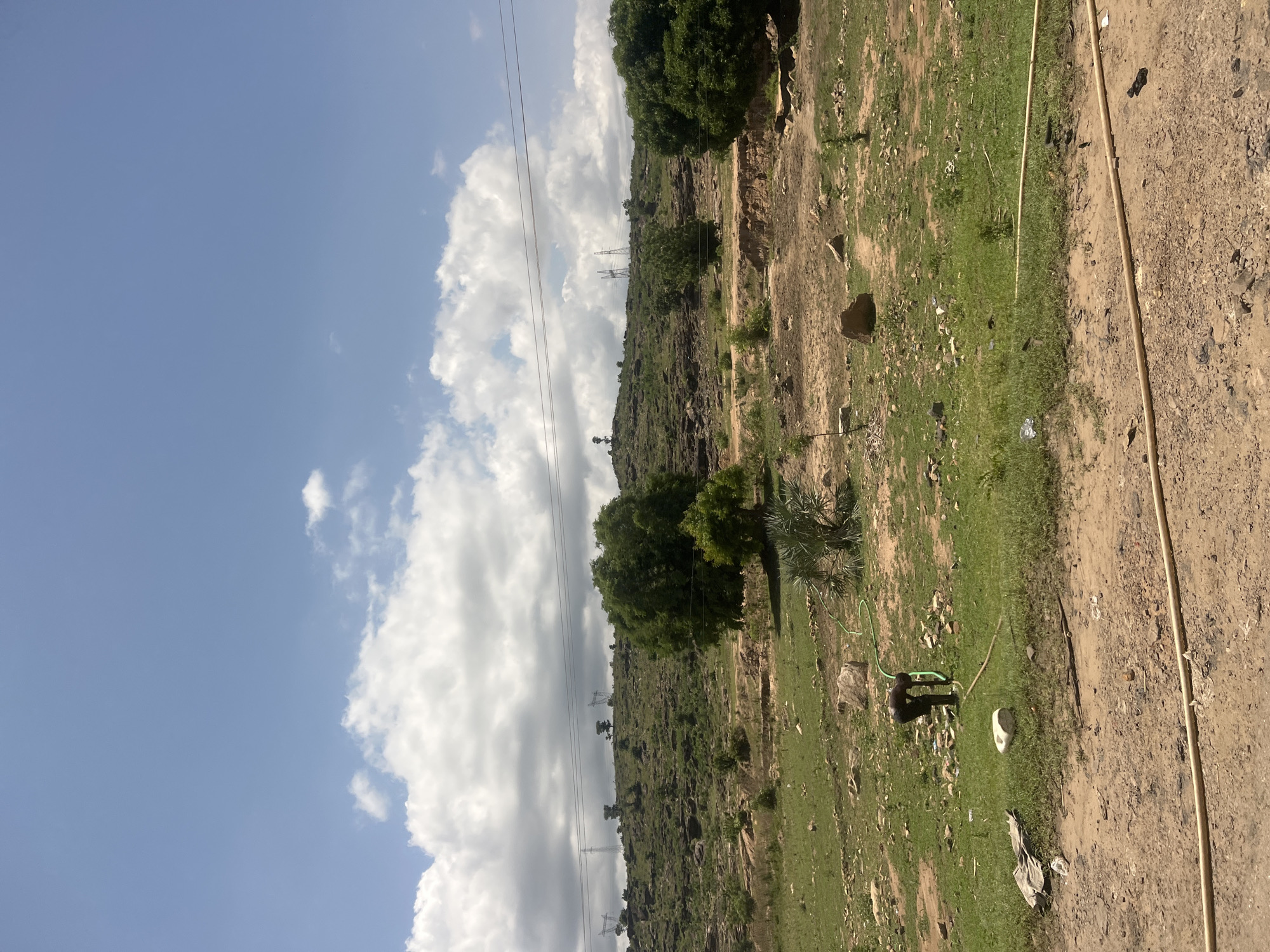 The image captures an expansive, semi-arid landscape under a partly cloudy sky, characteristic of an open area within Lafia, Nigeria. The upper portion of the frame features a clear blue sky interspersed with large, white cumulus clouds.
In the background, a range of rolling hills dominates the horizon. These hills are rocky, exhibiting exposed earth and sparse, scrubby green vegetation. Multiple high-voltage electricity transmission towers, characterized by their lattice steel structures, are visible along the hilltops, with power lines stretching horizontally across the scene.
The midground transitions into flatter terrain, a mix of dry, reddish-brown soil and patches of green grass. A distinct, eroded dirt embankment or small gully runs across the middle ground, indicating water flow or disturbance. Several trees with dense green foliage are scattered throughout this section, including a large, prominent tree on the far right.
The foreground consists primarily of a rough, reddish-brown dirt path or clearing, with scattered smaller rocks and some debris. Multiple lengths of light-colored, possibly tan or beige, hosing or piping lie horizontally across this area.
A lone individual, dark-skinned and dressed in dark clothing, is present in the lower-left midground. The person is bent over, facing away from the viewer, appearing to interact with a bright green garden hose that originates from the right and extends towards them. A white, possibly plastic, object rests on the ground near the individual. Additional scattered detritus, including what appears to be plastic waste and discarded fabric, is visible on the ground throughout the midground and foreground.
