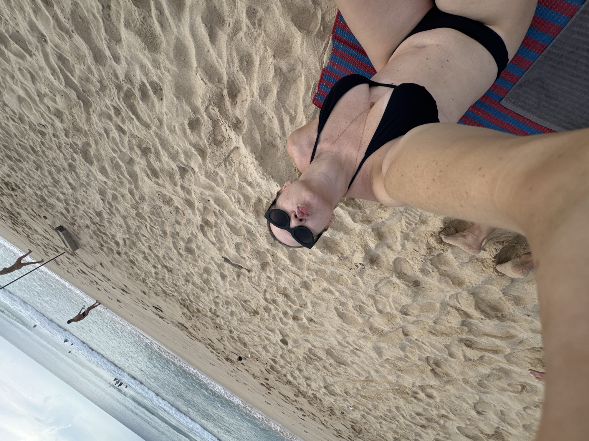 A woman in a black bikini is lying on a beach in Choeng Thale, Thailand, propped up on her elbows on a striped mat. She is wearing dark sunglasses and puckering her lips. Sand surrounds her, with footprints visible. In the background, the ocean and a few people are visible. The image appears to be a selfie taken from a low angle.