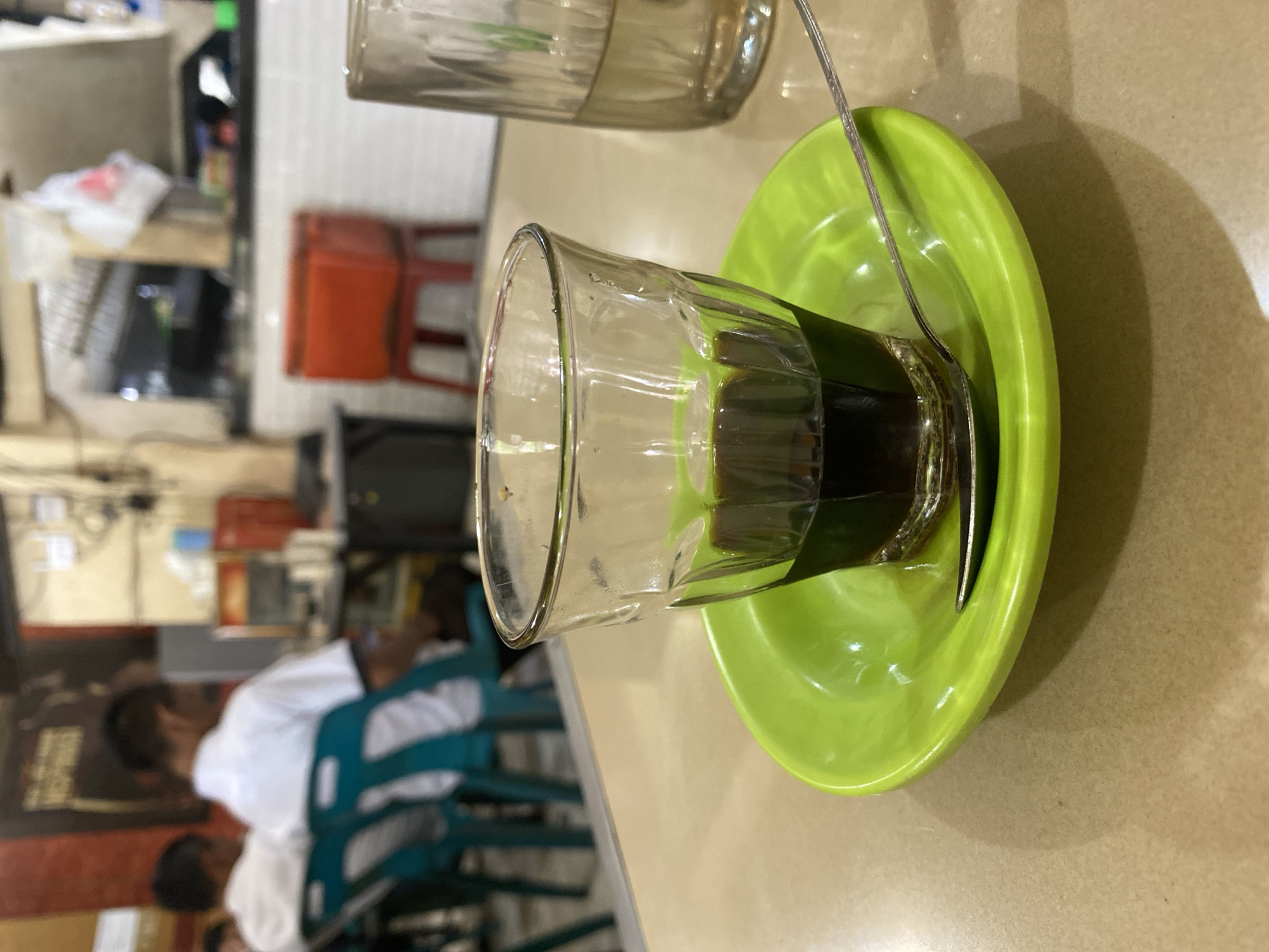 A close-up shot focuses on a small glass of dark coffee placed on a bright green saucer. A silver spoon rests in the saucer, partially submerged in the coffee. The glass is partially filled with liquid, and a dark leaf-like object is wrapped around its lower exterior. The coffee is dark and appears thick. The background is softly blurred, suggesting an indoor setting, possibly a cafe or restaurant. In the background, there are hints of other tables and chairs, as well as what appears to be a person wearing a white shirt and dark pants. There are also shelves with various items, including a red plastic container. The lighting suggests it might be daytime, but the interior setting makes it difficult to determine the exact time of day or weather. The overall impression is a simple, perhaps traditional, presentation of coffee. The scene is set in Lhokseumawe, Indonesia.
