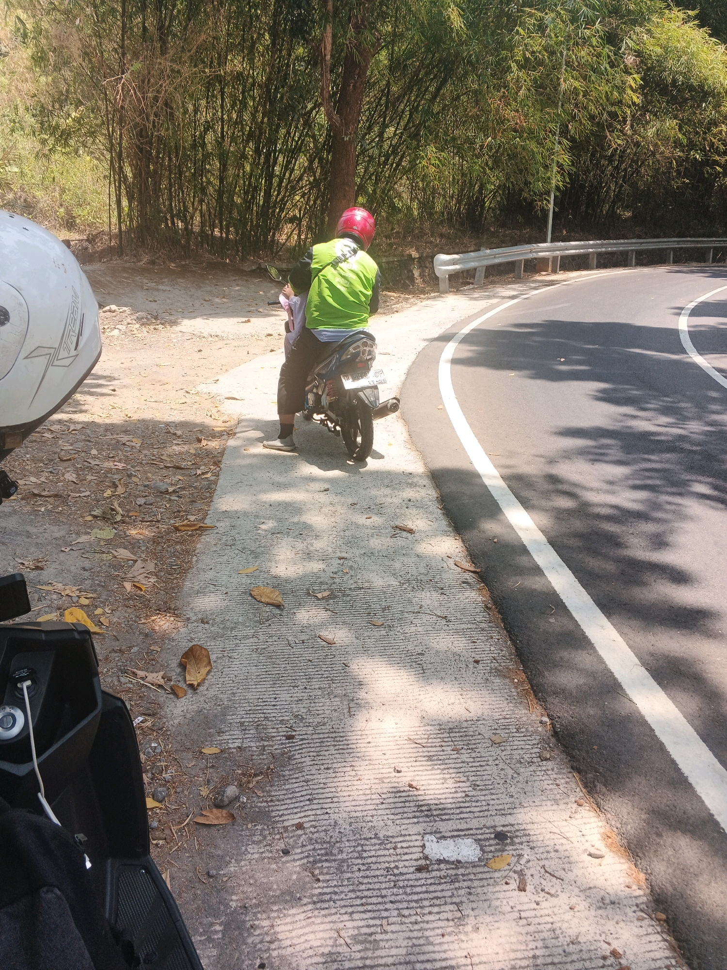 The image depicts a roadside scene in Semarang, Indonesia. In the foreground, the left side of a black scooter is visible, its storage compartment open and containing a white cable. To the right of the scooter, a person wearing a bright green jacket and a red helmet is seated on a motorcycle. The motorcycle is partially visible, showing a rear tire and exhaust pipe. The rider is facing away from the camera. Behind the motorcycle, a concrete path runs alongside a paved road. The road curves to the right, with a white line marking its edge. A metal guardrail is present along the side of the road. The background is dominated by a dense cluster of bamboo trees and other vegetation, indicating a natural or semi-natural environment. The ground is a mix of dirt, scattered leaves, and pebbles. Sunlight casts shadows across the scene, suggesting it is daytime.