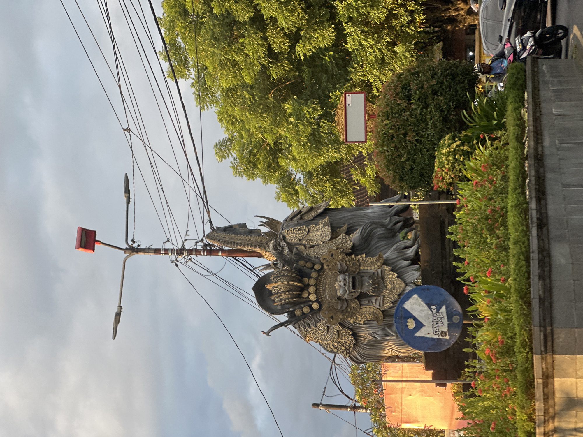 The image shows a street scene in Sukawati, Indonesia. In the foreground, there is a statue of a traditional Balinese deity. A blue and white direction sign is visible next to the statue, partially obscured by the statue. A street lamp and numerous overhead wires dominate the upper part of the frame. Lush greenery, including bushes and trees, lines the street. Further back, a parked motorbike, and car can be observed. A sign with a white face is mounted on a pole. The overall impression is one of a typical Balinese street, blending traditional art with urban infrastructure.