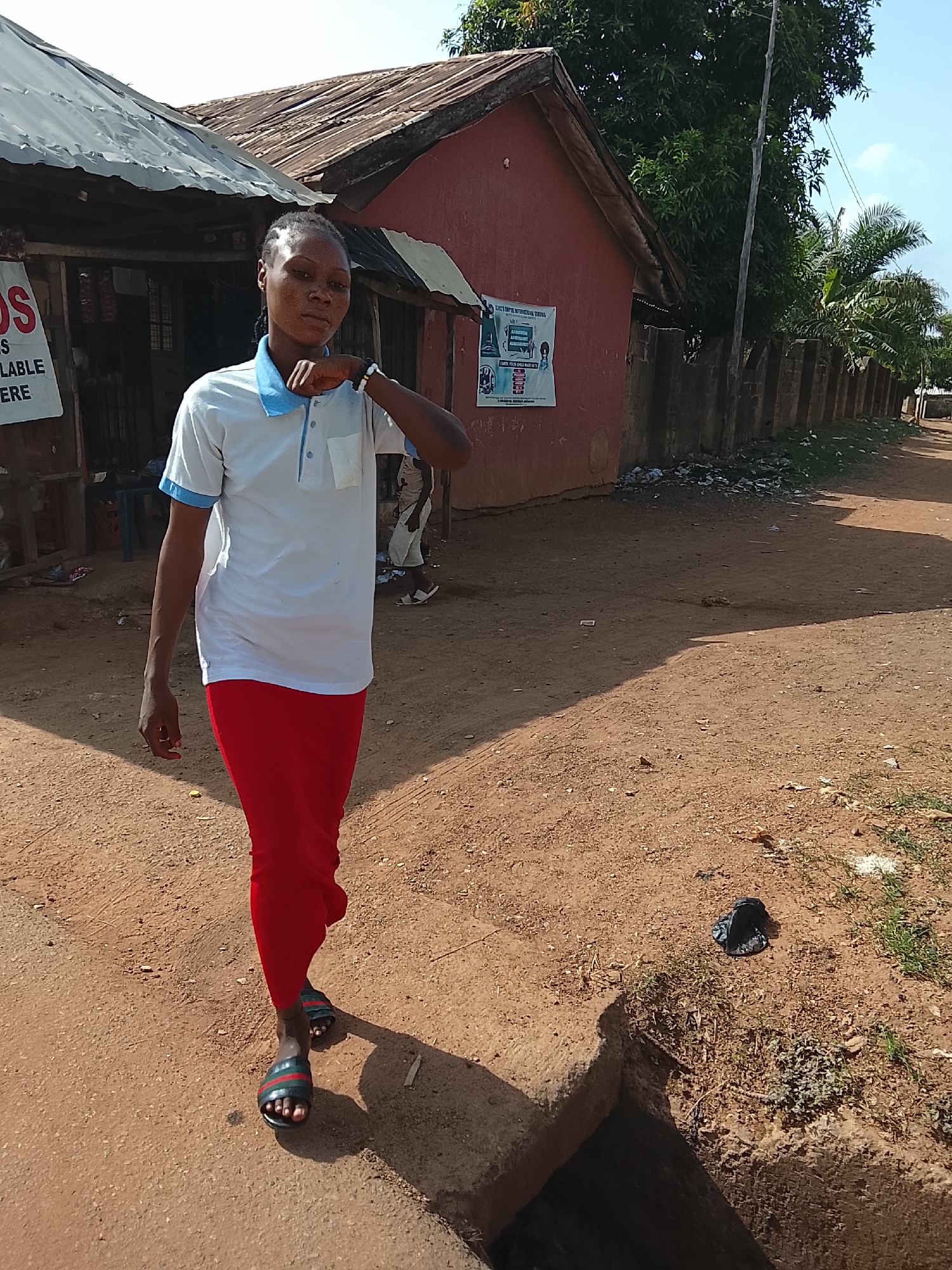 A young adult with dark skin and braided hair stands on an unpaved, reddish-brown dirt path, posing for a photograph. The subject is wearing a white short-sleeved polo shirt with light blue trim on the collar and sleeves, and bright red trousers. Dark, striped slides are visible on their feet. Their right hand is casually raised to their chin or neck area, and they are looking directly at the camera.
The scene appears to be an everyday setting in Akwanga, Nigeria, under bright, clear daylight, casting distinct shadows. To the left, a simple building with a corrugated metal roof, possibly a small shop, features a partial white sign with red text that reads "DS", "S", "IS", "AVAILABLE", and "HERE". In the mid-ground on the right, another building, painted reddish-brown, also has a corrugated metal roof. A white and blue poster is affixed to its wall, advertising "LET'S EMPIRE INTERNATIONAL SCHOOLS" and details about "ADMISSION", "SUMMER CLASSES", and "ENROLL YOUR CHILD".
Further along the path to the right, a low concrete block fence runs parallel to the road, with lush green trees, including palm trees and other foliage, visible behind it. The ground is mostly dry dirt with scattered small patches of grass and some litter, including a crumpled black plastic bag near a concrete drainage ditch in the foreground. The overall impression is a vibrant, sunlit candid shot in a local community environment.