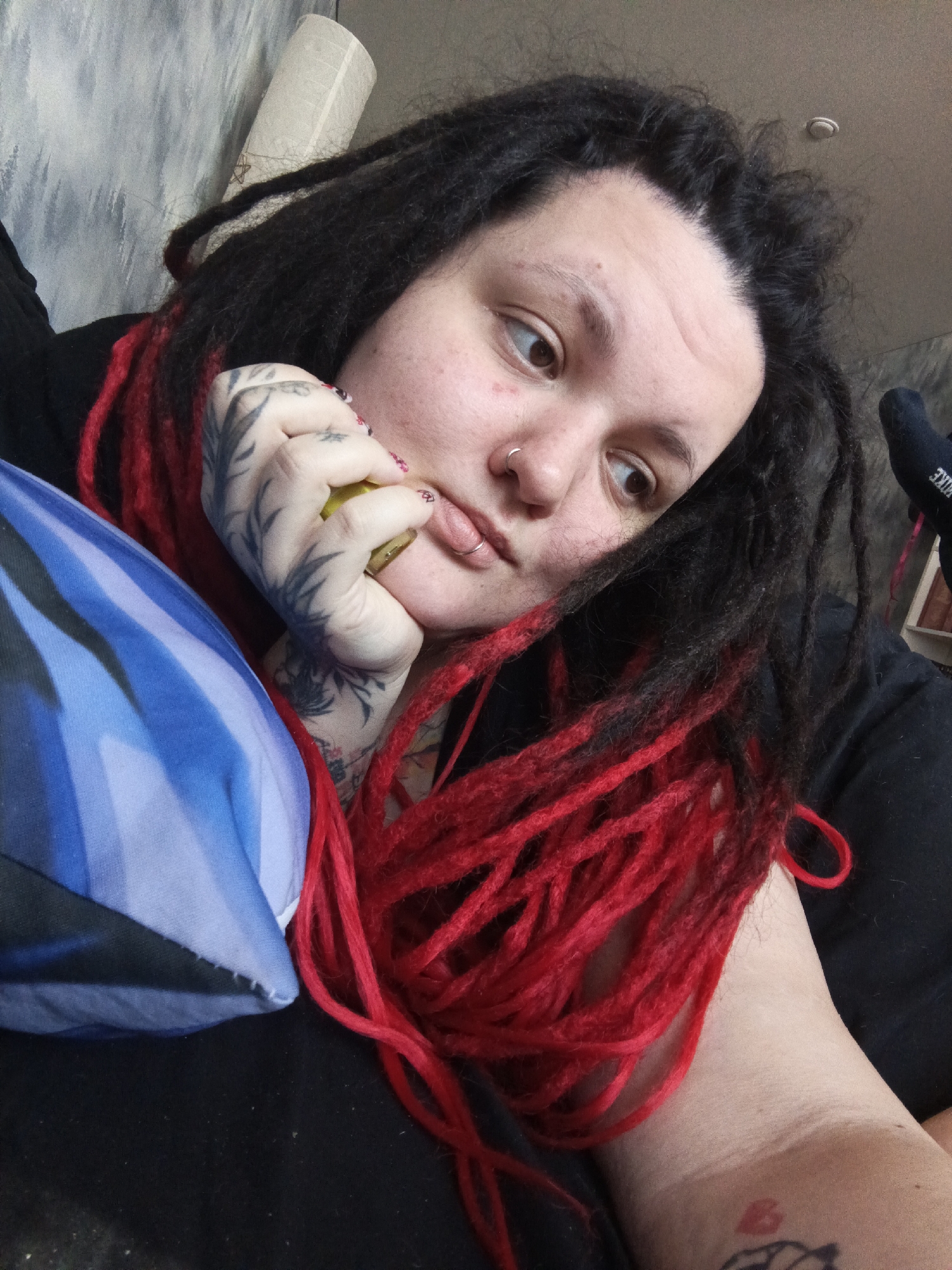 The image displays a close-up portrait of an adult female with a light complexion, dark dreadlocks, and prominent sections of bright red synthetic dreadlocks. She has brown eyes, a silver hoop nose ring in her left nostril, and a silver hoop lip ring on her lower lip. Her left hand, adorned with extensive black ink tattoos featuring a foliage-like design, is positioned near her chin, holding a small, yellowish-green object, possibly a pickle, near her mouth. Remnants of chipped red nail polish are visible on her fingernails. She is wearing a dark, possibly black, long-sleeved garment. A red heart-shaped tattoo is visible on her inner right forearm.
To her left, a pillow or draped fabric with horizontal blue and light blue stripes is present. In the background, a light-colored wall with a subtle textured finish is visible, with a rolled-up object featuring a star pattern leaning against it in the upper left. A light-colored ceiling with a small, circular fixture (possibly a smoke detector or light) is in the upper center. In the upper right, a black sock with "NIKE" written in white is visible. The scene appears to be an indoor, likely domestic, setting. The image does not contain visual evidence to confirm its specific location within the city of Chaikovsky, Russia.