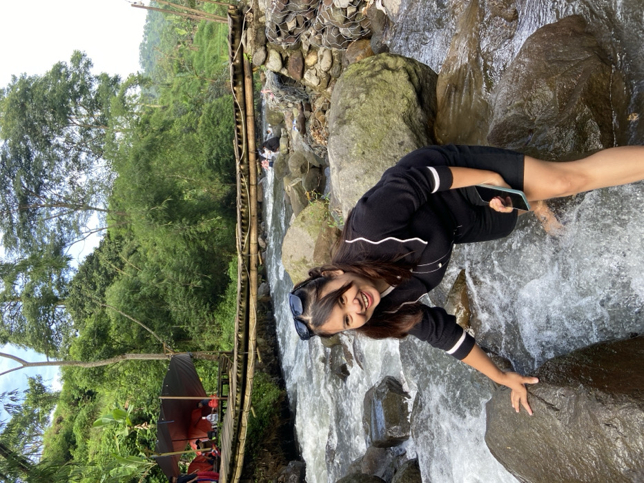 A young woman kneels by a rocky stream, her hand resting on a large stone. She wears a black, long-sleeved shirt with white stripes on the sleeves, and black shorts. Sunglasses rest on her head, and she smiles at the camera. A phone is in her hand. Lush green trees surround the area, with a wooden structure and red umbrellas visible in the background. The water rushes over the rocks, creating a dynamic and natural setting.