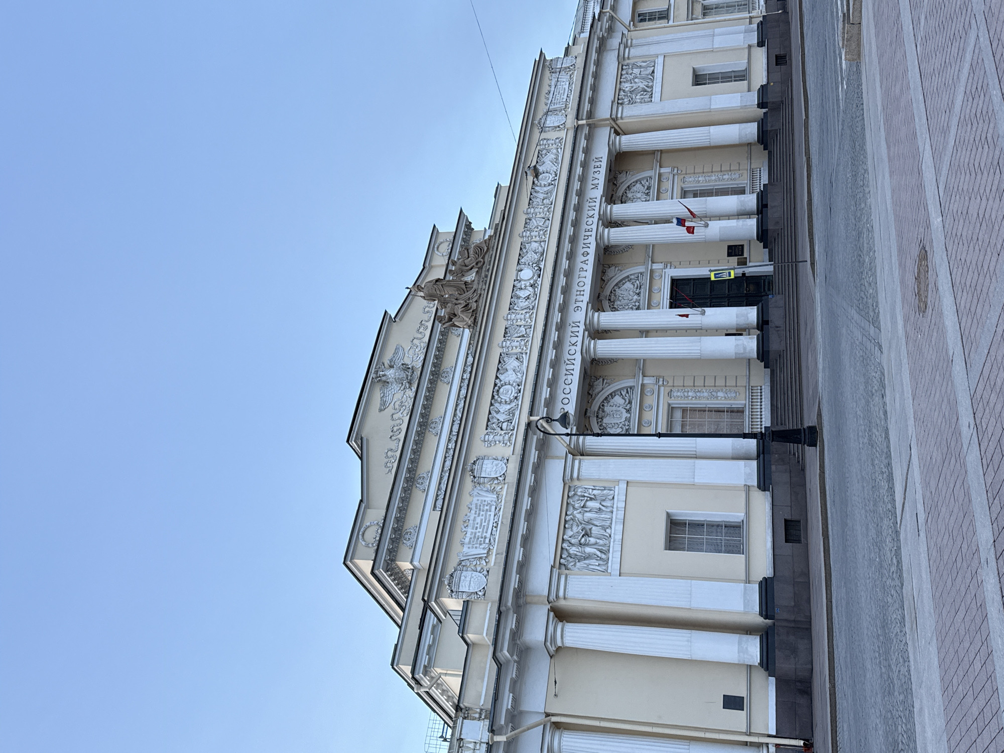 The image shows the facade of the Russian Museum of Ethnography in Saint Petersburg, Russia. The building is neoclassical in style, with a pale yellow exterior, white columns, and detailed sculptural friezes. The text on the building reads "РОССИЙСКИЙ ЭТНОГРАФИЧЕСКИЙ МУЗЕЙ" (Russian Museum of Ethnography). The museum's entrance is visible, with flags of Russia and other countries on either side. A street runs alongside the building, with a sidewalk paved in red and gray bricks. The sky above is clear and blue.