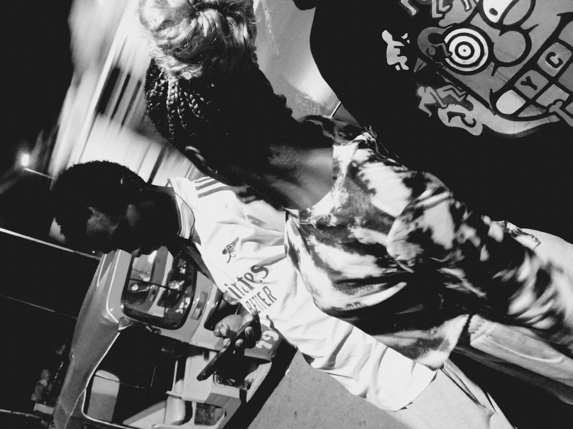A black and white photograph depicts two individuals next to a vehicle in what appears to be the city of Bukuru, Nigeria. One individual, presumably male, is wearing a light-colored Arsenal jersey with "Emirates Better" printed on it, and is holding a dark object (possibly a phone) in his hand. The other individual has braided hair and is partially obscured, wearing a shirt with a graphic design. The vehicle is a van. The overall scene is grainy and appears to be taken at night or in low light. Evidence indicating Bukuru, Nigeria, is not apparent in the provided image.