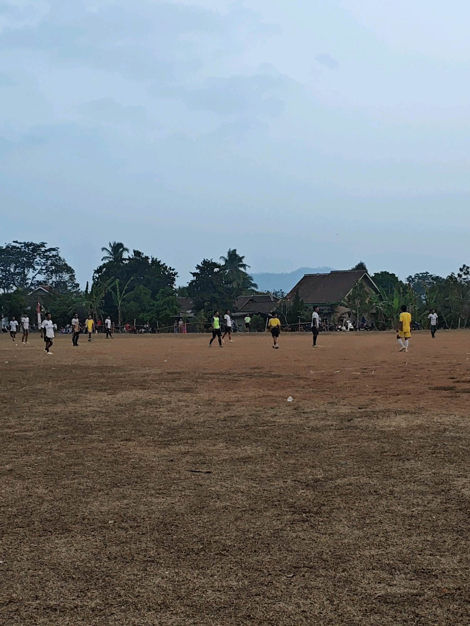 This is a wide, horizontal, medium-shot, eye-level photograph of a soccer game taking place on a dry, brown dirt field under a hazy, overcast sky. The field is sparsely covered with dry grass and debris. In the background, there are several trees and some buildings, including houses with brown roofs and a larger, darker building. Some people are visible in the distance, appearing to be spectators.
In the foreground and midground, several individuals, presumably players, are scattered across the field. They are wearing various colored shirts and shorts, including white, yellow, and dark colors. Some players are standing, while others appear to be in motion, suggesting an ongoing game. The players' actions and interactions are not clearly discernible due to the distance and resolution. The overall impression is of a casual outdoor sporting event in a rural or semi-rural setting.