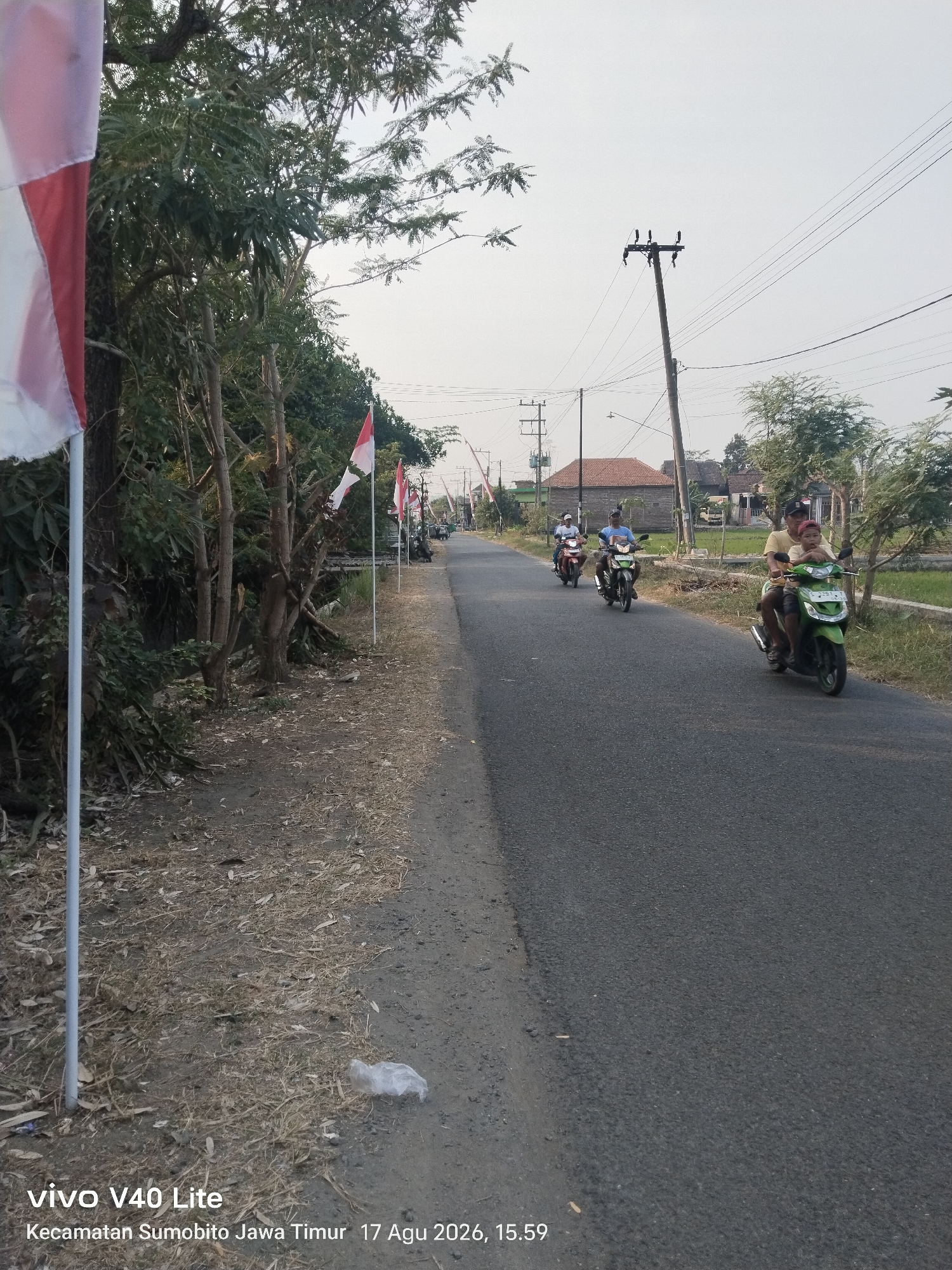 The image depicts a street scene in Mlaras, Indonesia. On the left side of the paved road, a white flag pole with a red and white flag is visible, partially obscured by trees. Further down the road on the left, several more red and white flags are planted along the roadside. The road itself is asphalt and appears to be a rural or semi-rural area, with vegetation and some houses visible in the background. A power pole with electrical wires stands on the right side of the road. Several people are riding motorcycles on the road. In the foreground on the right, a green motorcycle carries two individuals. Another motorcycle with two riders is further ahead, and a third motorcycle with a single rider is in the distance. The sky is overcast. The bottom of the image contains a watermark displaying "vivo V40 Lite" and text indicating "Kecamatan Sumobito Jawa Timur 17 Agu 2026, 15.59".