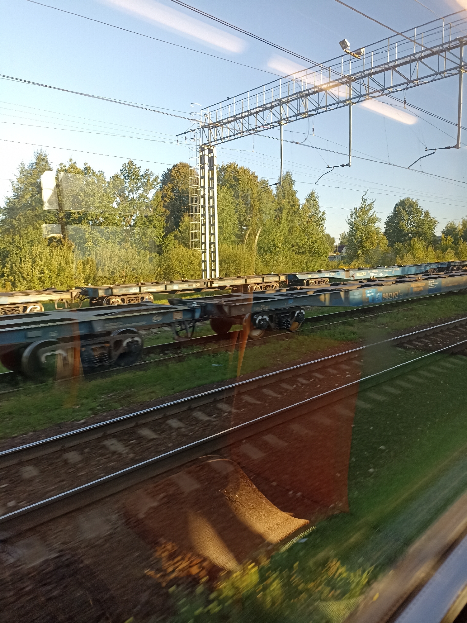The image, taken from a moving train, captures a view of freight train cars on parallel tracks under a clear blue sky. The train cars are long, flat, and blue, with visible wheels and undercarriages. Some of the cars have markings that appear to be serial numbers, such as "94143492." In the background, a line of lush green trees can be seen bordering the railway. Overhead, complex electrical infrastructure for the train, including wires and support structures, is visible against the sky. The foreground shows the train tracks themselves and some reflections on the window, suggesting the image was taken from inside another train. The scene appears to be in a rural or semi-rural area, consistent with the location of Stolbovaya, Russia, a town known for its railway junction. There are no people visible in the image.